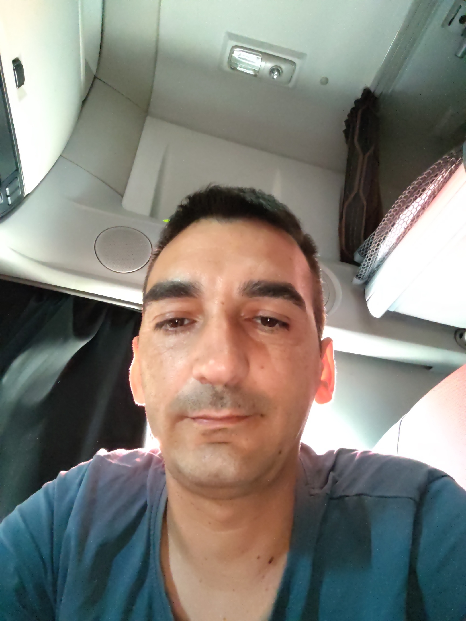The image is a selfie taken inside a vehicle cabin, likely a truck, looking upwards at the subject. The man, with dark hair and a mustache, is wearing a teal t-shirt. The cabin interior is visible, featuring white paneling, a light fixture on the ceiling, and dark curtains. The lighting is bright, with some of it appearing to come from above and behind the subject, casting a warm glow on his ear. There are no discernible landmarks or elements that would specifically place the location in Authume, France. The focus is on the subject's face and upper torso, with the cabin environment serving as a backdrop.