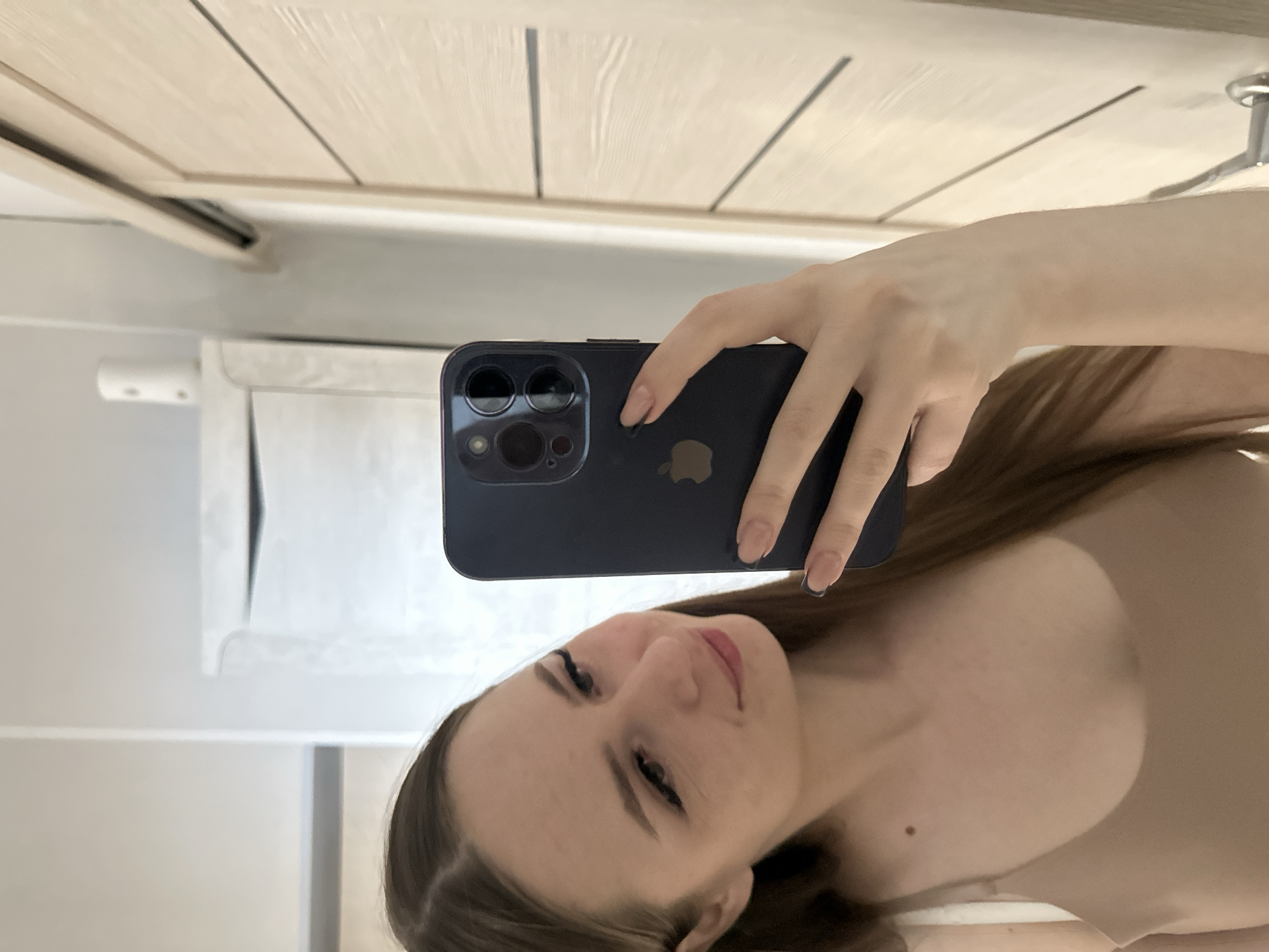 A woman is taking a selfie indoors, holding a dark-colored iPhone in her right hand. The phone is positioned to capture her face, which is slightly tilted towards the camera. Her hair is light brown, and she is wearing a neutral-colored top. The background includes a white wall with a wooden door at the top, and a partially visible shelf or ledge. The overall environment appears to be a residential interior, though the specific location and city are unknown.