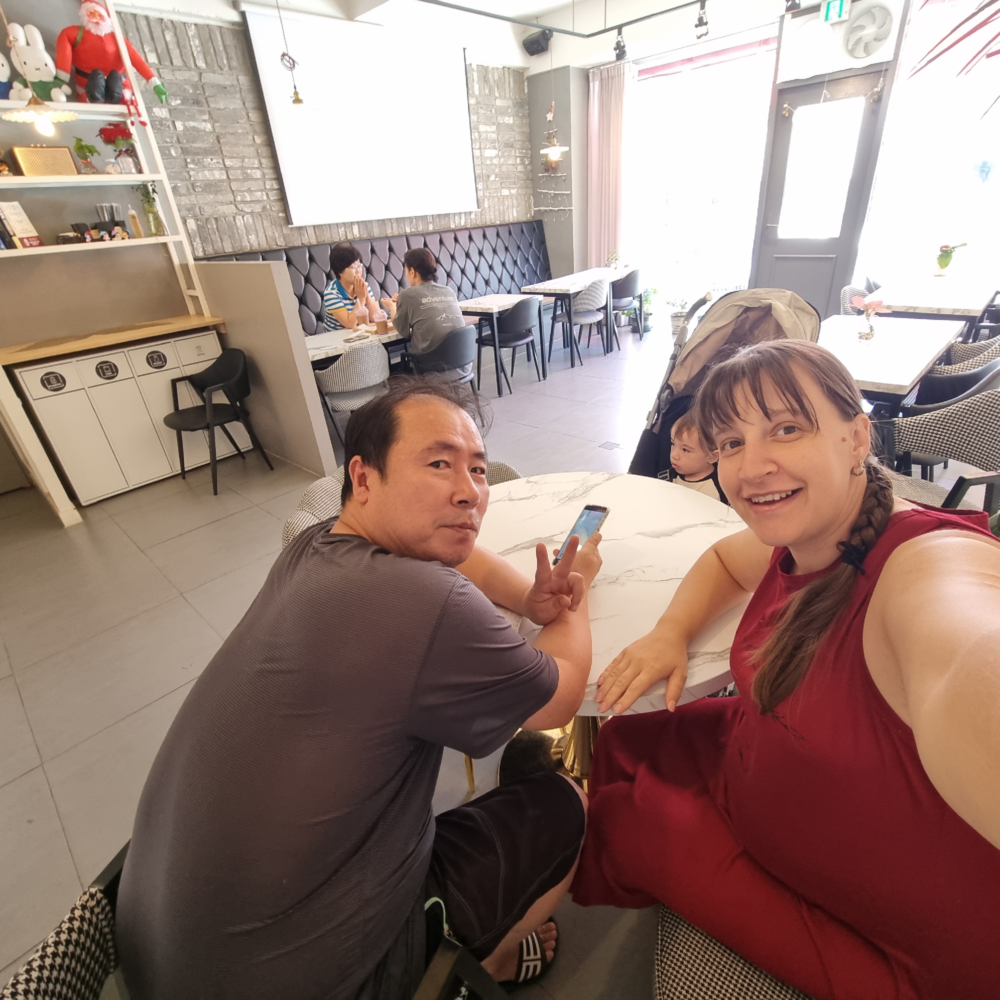 The image captures a scene inside a brightly lit cafe, likely located in Dunnae-myeon, South Korea. In the foreground, a man in a grey t-shirt and black shorts is seated at a marble-topped table, holding a smartphone and making a peace sign with his fingers. Next to him, a woman in a red sleeveless dress is smiling broadly at the camera, her braided hair visible. A young child with a concerned expression is seated in a stroller behind the woman. In the background, another table is occupied by two women, one in a blue and white striped shirt and the other in a grey shirt, engaged in conversation. The cafe's interior features grey brick walls, a black tufted banquette, and modern furniture. A tall shelf unit displays various items, including a Santa Claus doll and other decorative objects. The overall atmosphere appears casual and relaxed, suggesting a family outing or a casual meetup at a local establishment.