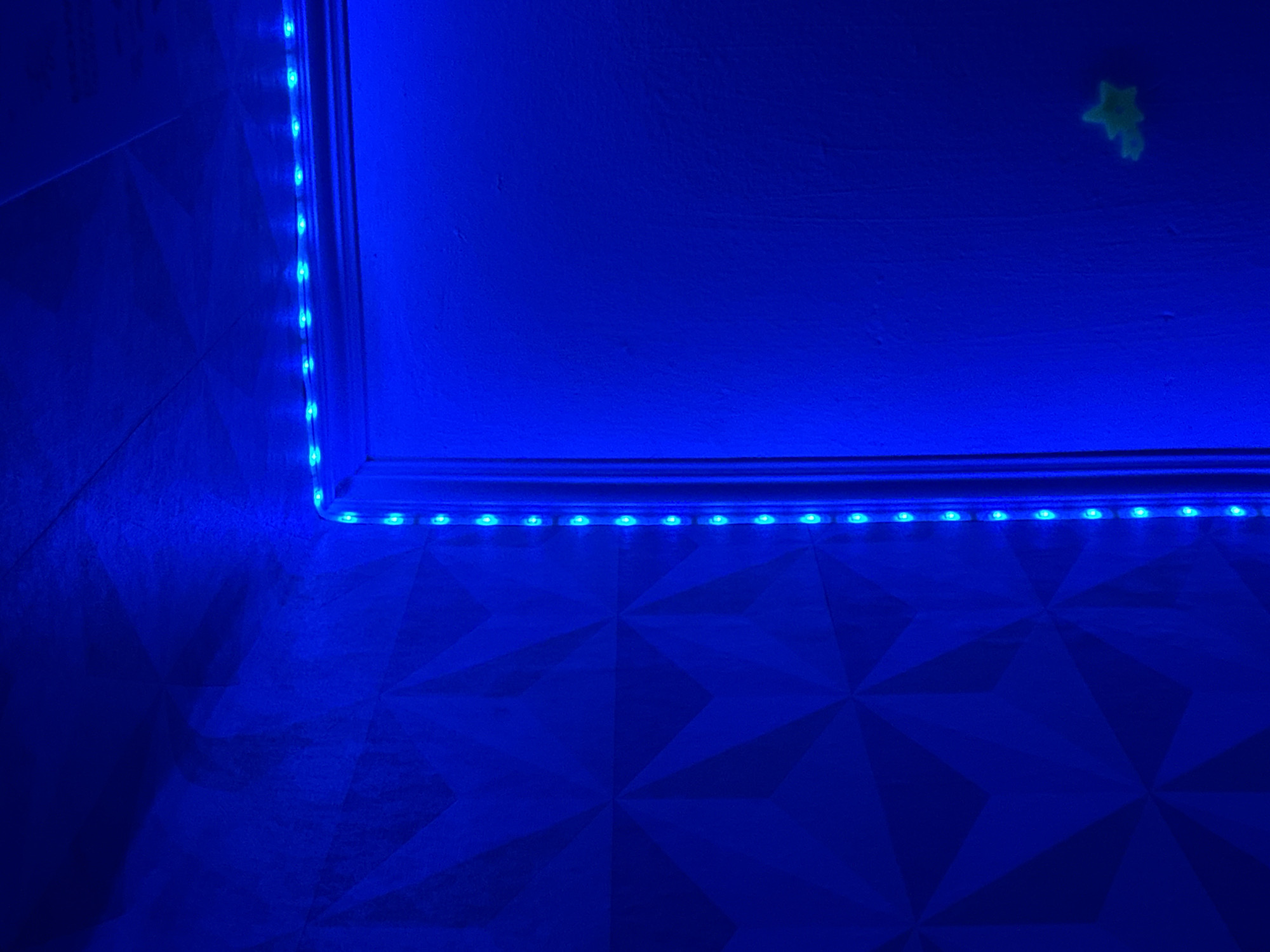 A low-angle shot shows a room corner illuminated by blue LED strip lights. The lights run along the top edges of two walls, creating a blue glow across the surfaces. The walls and floor have a geometric pattern, with a repeated star-like design. The blue light enhances this pattern, casting shadows that accentuate the three-dimensional texture of the walls and floor. A small, faintly glowing star-shaped object is visible on the upper wall. The overall impression is one of an artificially lit interior, possibly a bedroom or lounge. The location of Chaikovsky, Russia is not apparent from the image.