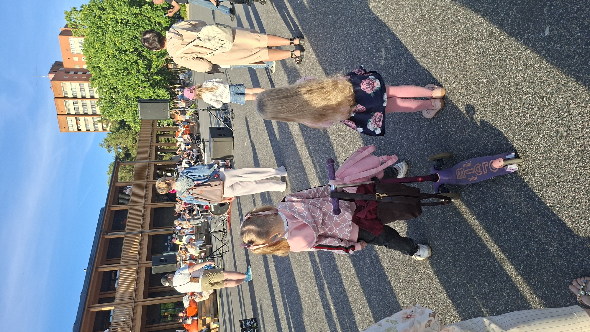 In Tallinn, Estonia, an outdoor gathering is underway with a stage and seating area visible in the background. The scene is populated by numerous individuals, primarily adults and children, milling about or seated. In the foreground, two young girls are prominently featured. One, facing away from the camera, wears a pink hooded jacket with a repeating logo pattern, dark pants, and has blonde hair styled in pigtails with pink bows. She stands next to a purple scooter with a bag and another pink backpack attached. The second girl, also facing away, has long blonde hair and wears a dark dress with a floral pattern and pink tights. Behind them, other attendees are visible, including a woman in a light-colored hooded jacket and another in a beige jumpsuit carrying a backpack. Speakers and audio equipment suggest a performance or presentation is taking place on the stage, which is situated in front of a wooden building with multiple levels and balconies, surrounded by trees and a tall brick building in the distance. The ground is paved asphalt, and shadows are cast by the low sun, indicating it is late afternoon or early evening.