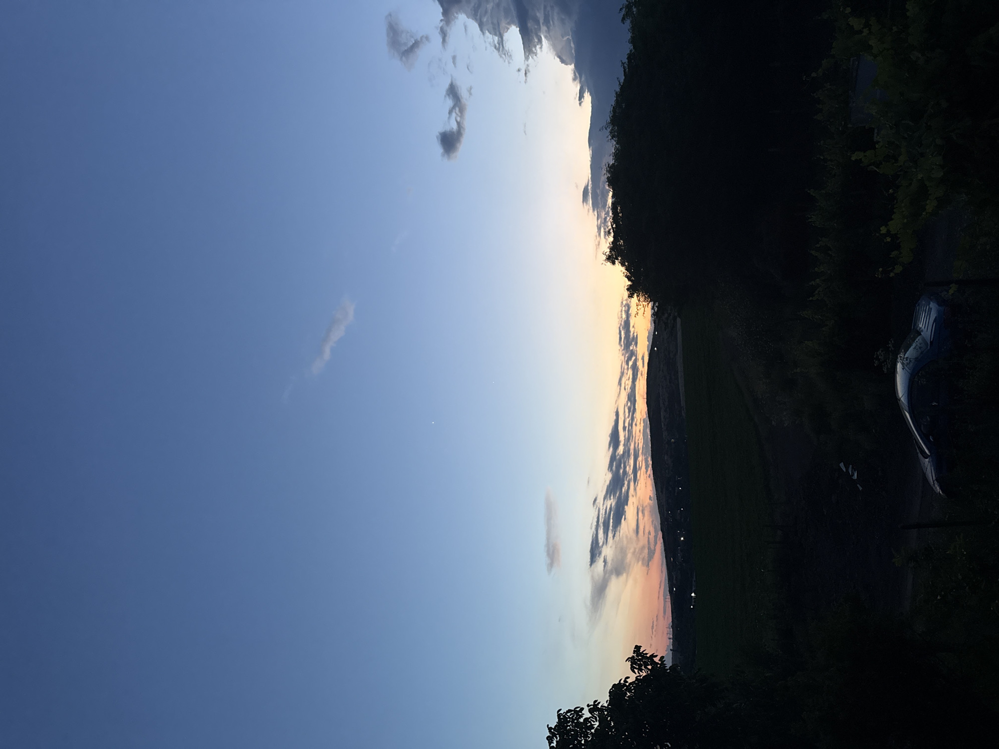 The image depicts a twilight scene, likely captured in or near Burgas, Bulgaria, given the context. The sky is a gradient of blues, fading to a warm orange and pink at the horizon where it meets a silhouetted hill. Wispy clouds are scattered across the sky, some catching the last light of the setting sun. In the foreground, dark silhouettes of trees and foliage frame the view. A dark-colored car is parked in the lower left portion of the frame, partially obscured by shadows. A dark, cultivated field is visible in the mid-ground, with distant lights dotting the hillside. The overall impression is one of a peaceful, rural evening on the outskirts of a city.