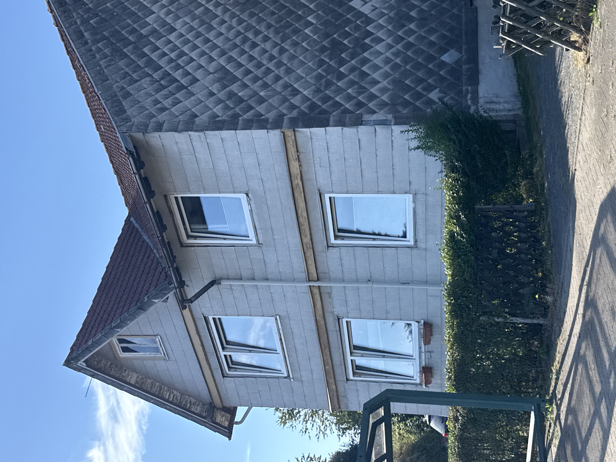 The image captures the exterior of a two-story house in Bad Sachsa, Germany, under a bright blue sky with scattered clouds. The house features a pitched, dark red tiled roof. The upper part of the front facade is covered in light gray horizontal siding, with a gable end showing a small window and peeling white paint on the trim. A dark gutter system is visible along the roofline. The main facade has two windows on the upper floor and two on the lower floor, all with white frames. Above the lower windows, there is a horizontal wooden beam. The right side of the house is clad in a dark gray, diamond-patterned material. In front of the house, a dense green hedge runs along a low, dark green fence. To the left, part of a bus stop shelter with a dark green frame is visible, and a red car can be seen parked behind the hedge. The ground in front of the hedge is a paved area with a textured surface, casting distinct shadows. A rustic wooden fence is positioned on the right side of the image, near the base of the house.