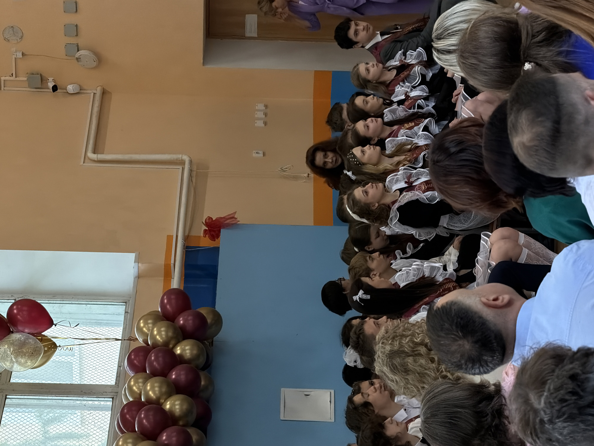 The image depicts a graduation ceremony, likely in a school or auditorium setting, filled with students dressed in traditional graduation attire. It's suspected to be taken in the city of Podolsk, Russia.
People: A large group of people is present, predominantly young students. The students are wearing dark dresses with white lace collars and cuffs, typical of a graduation ceremony in Russia. Many are wearing sashes that say "выпускник" (graduate). Some adults are visible as well, but they are in the background or partially obscured. The general focus of the image is on the students.
Subjects: The subjects primarily include the students, likely high school graduates, and the event itself—a graduation ceremony.
Objects: Balloons are visible, with a mix of red, gold, and clear colors. They are situated next to a window. The setting appears to be indoors.
Actions: The students are seated, facing forward, and appear to be listening or watching an event. There are likely interactions happening, such as conversations.
Interactions: The primary interaction is between the students and whoever is leading or speaking at the ceremony.
Environment: The indoor environment has walls that are painted in two tones: light yellow on the upper part and blue on the lower part. The natural light is visible. The setting seems to be a standard school or auditorium.