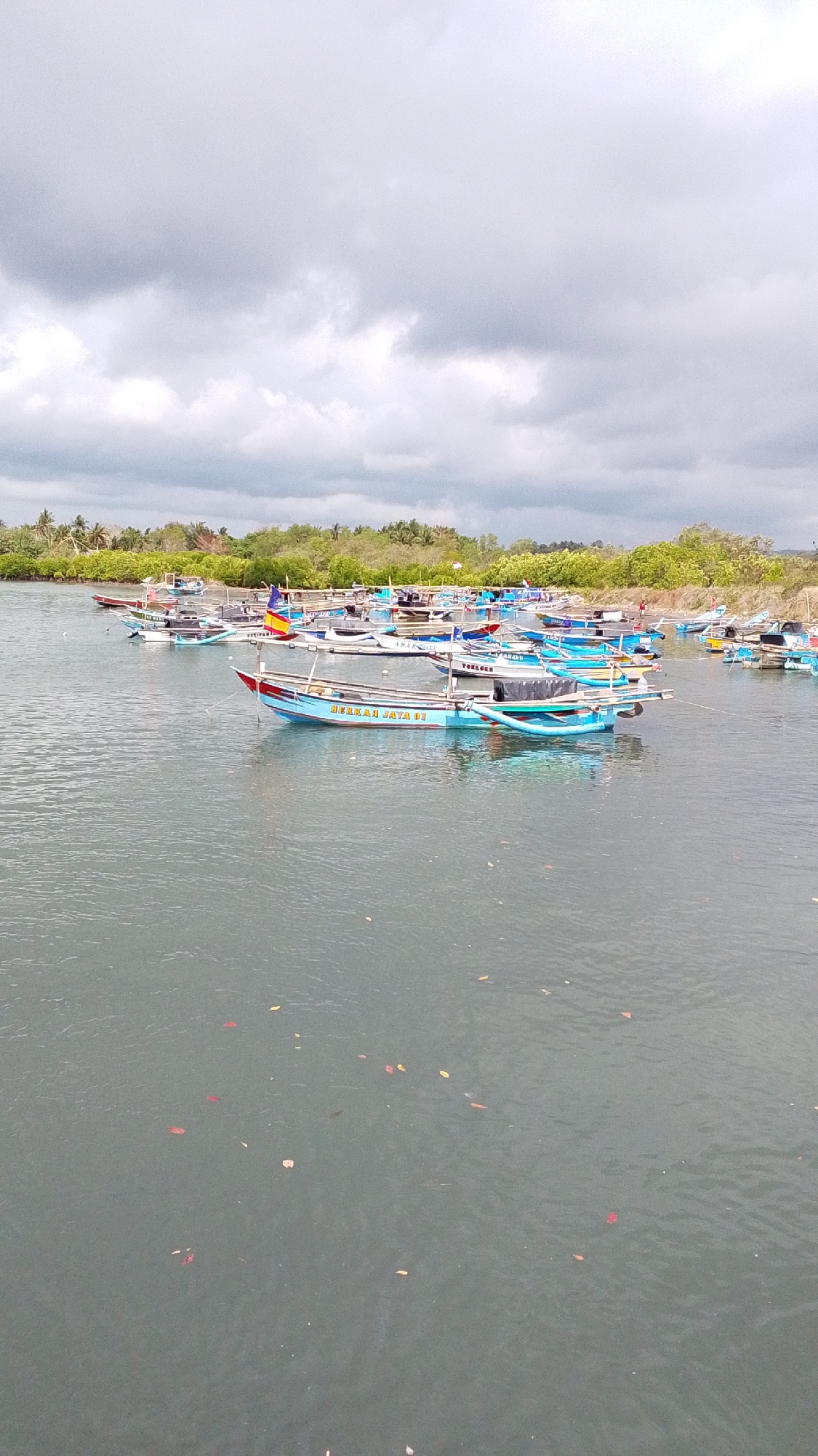 The image depicts a fleet of small fishing boats docked in a body of water, likely a river or a bay, with a lush green coastline in the background under a cloudy sky. The boats are of various sizes and colors, with some displaying flags. One prominent boat in the foreground has the text "BERKAH JAYA 01" written on its side in yellow lettering. The water is calm, with a few fallen leaves floating on the surface. The scene appears to be a typical day at a fishing village in Desa Karangjaladri, Indonesia, a coastal village known for its fishing activities and located in the Jarat district of the Pemalang Regency, Central Java.