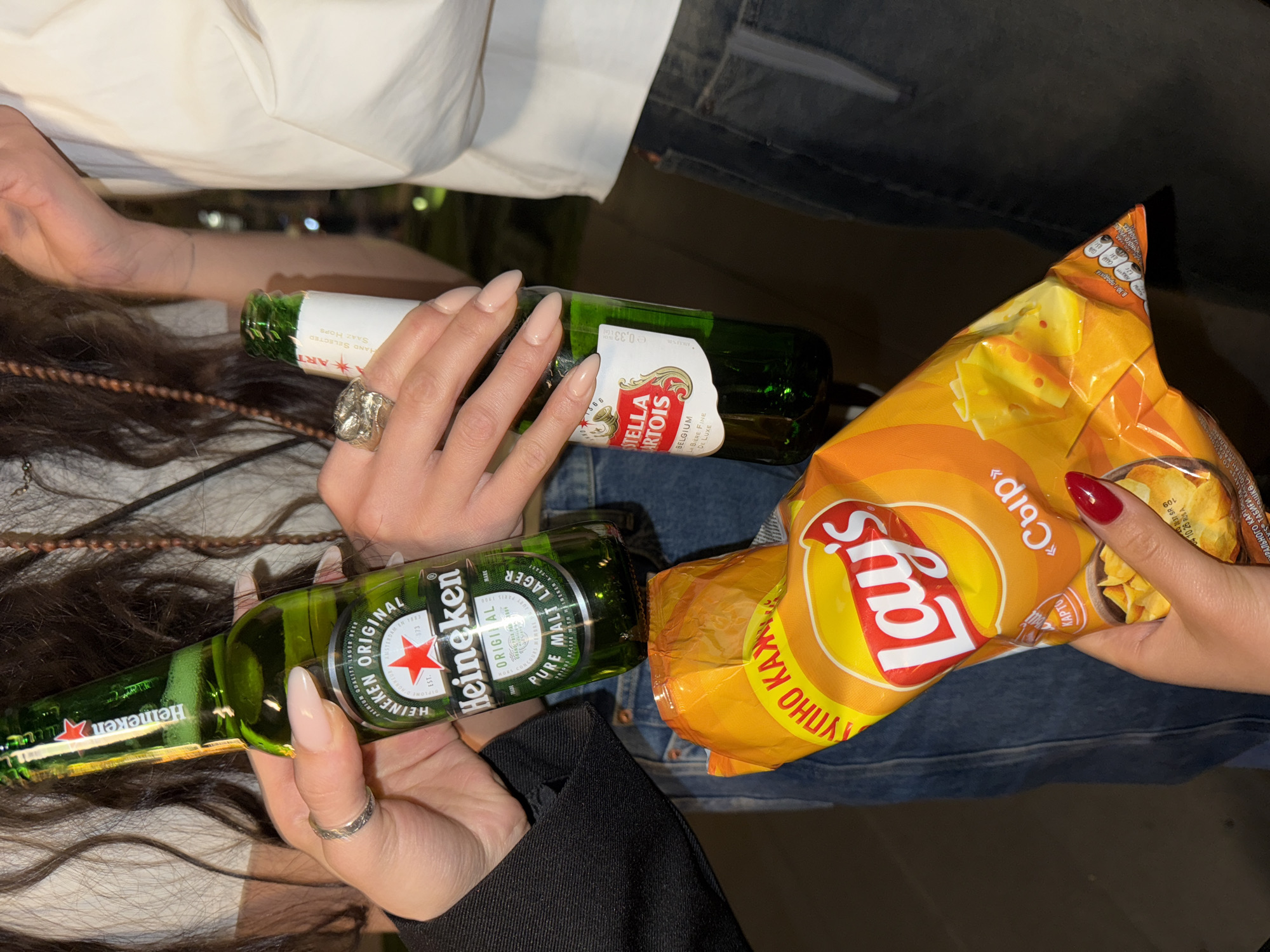 The image presents a top-down view of a gathering where three people are holding snacks and drinks.
One person is holding a bottle of Heineken, with their hand displaying a silver ring. Another individual is holding a bottle of Stella Artois, their hand adorned with a large silver ring. The third person holds a bag of Lay's "Cheese" flavored chips, with their hand featuring red nail polish. All three are close together, indicating a shared space or activity.
The clothing worn by the people consists of a white shirt, black jacket, and blue jeans.
The image provides no information about the city, Yerevan, Armenia. The location cannot be determined from this picture.