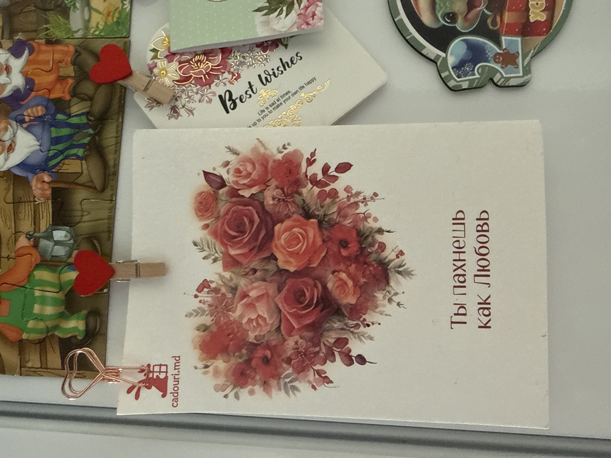 The image displays a close-up view of a refrigerator door adorned with various decorative items. Prominently featured is a postcard with a heart-shaped floral arrangement in shades of red and pink. Below the floral design, text in Russian reads "Ты пахнешь как Любовь" which translates to "You smell like love." To the left of the postcard, a rose gold heart-shaped paper clip holds a small image of a rabbit with a gift. To the right of the postcard, another postcard with the words "Best wishes" is partially visible, featuring floral embellishments. At the bottom right, a circular magnet with the number "2" and a cartoon character holding a gift box is attached. The background shows a puzzle of gnomes and a garden scene. The items are likely displayed on a refrigerator door in a residential setting. Given that one of the visible postcards has "cadouri.md" printed on it, which is a website for gift delivery in Moldova, it's possible this scene is located in Chișinău, Moldova.