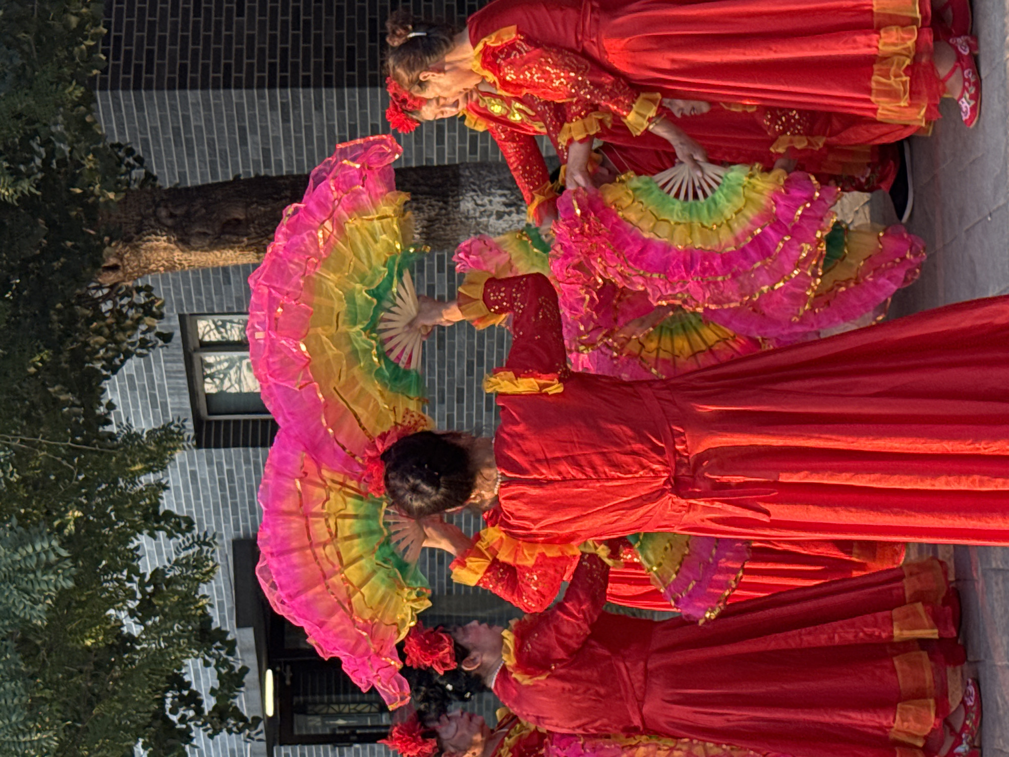 The image depicts a group of women in vibrant red traditional Chinese dresses, adorned with yellow trim and gold sequins, performing a fan dance outdoors. The fans are large, circular, and richly colored in pink, yellow, and green, with gold trim. The women are positioned in a semi-circle, with some facing away from the camera and others towards it. Their hair is styled up, and some wear red floral hair accessories. The background features a textured grey brick building with a dark-colored roofline and windows. A mature tree with rough bark stands behind the dancers, its branches and leaves partially obscuring the building. The lighting suggests it is late afternoon or early morning, with a warm glow illuminating the scene. The ground appears to be paved with stone or brick. The overall atmosphere is festive and cultural, possibly at a public gathering or performance in Jinzhou, China.
