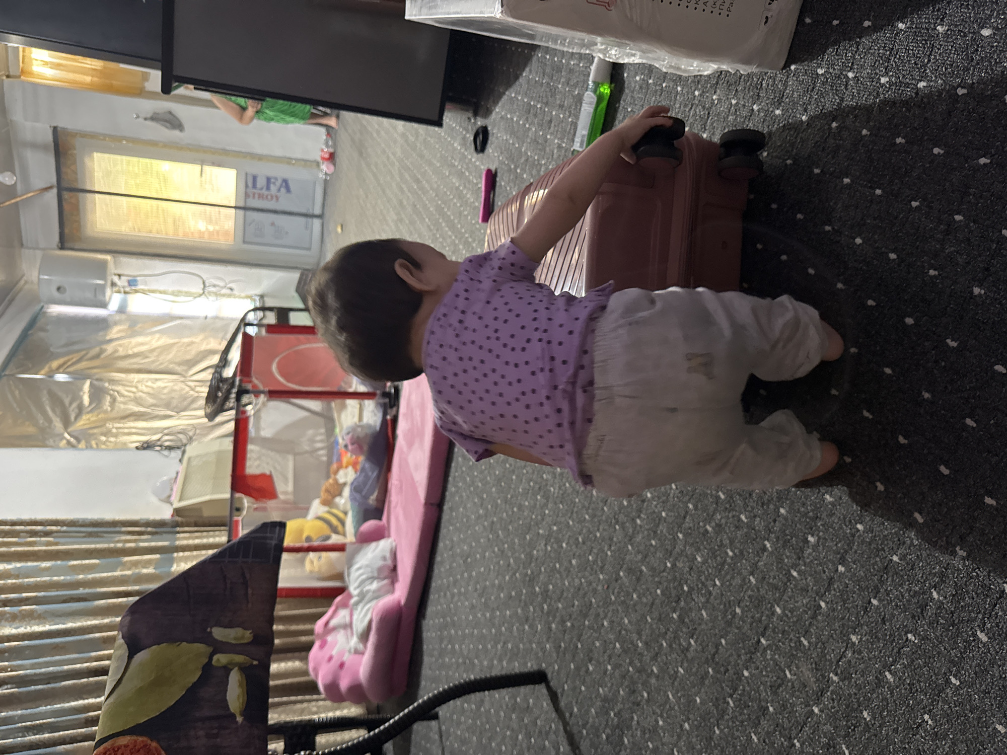 The image captures an indoor scene with a young child, viewed from the back, reaching for a pink suitcase on a dark, patterned carpet. The child is wearing a purple polka-dot t-shirt and light-colored pants. In the background, a pink baby crib with a mattress is visible, along with a plush pink toy chair. Further back, a woman in a green dress stands near a doorway with a yellow-tinted window. The room appears to be a residential space, possibly in Chardara, Uzbekistan, given the context provided. Other objects in the room include what appears to be a water heater on the wall, foil insulation, and various other items that suggest a work-in-progress or storage area. The lighting is dim, with a light source emanating from the doorway.