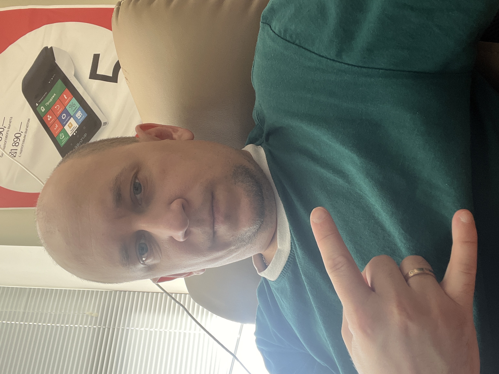 A man is pictured in a close-up selfie, seated in a chair. He has a shaved head and light-colored skin, with a slight goatee. He is wearing a dark green t-shirt with a white collar visible. His left hand is raised, with his index and pinky fingers extended in a rock-and-roll gesture, and he is wearing a gold band on his ring finger. In the background, there is a large advertisement featuring a handheld payment terminal and text in Russian. To the left of the frame, vertical blinds are visible. The overall scene suggests an indoor setting, possibly an office or retail space. There is no specific indication of the city of Omsk, Russia within the image.