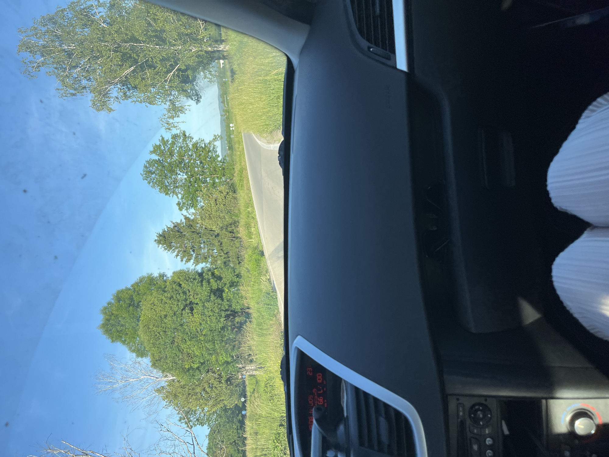 The image shows a view from inside a car, likely from the driver's seat. Outside the car, there's a road curving through a landscape of green fields and trees under a blue sky. The dashboard of the car is visible, with vents and controls. The location is Unknown, Unknown.