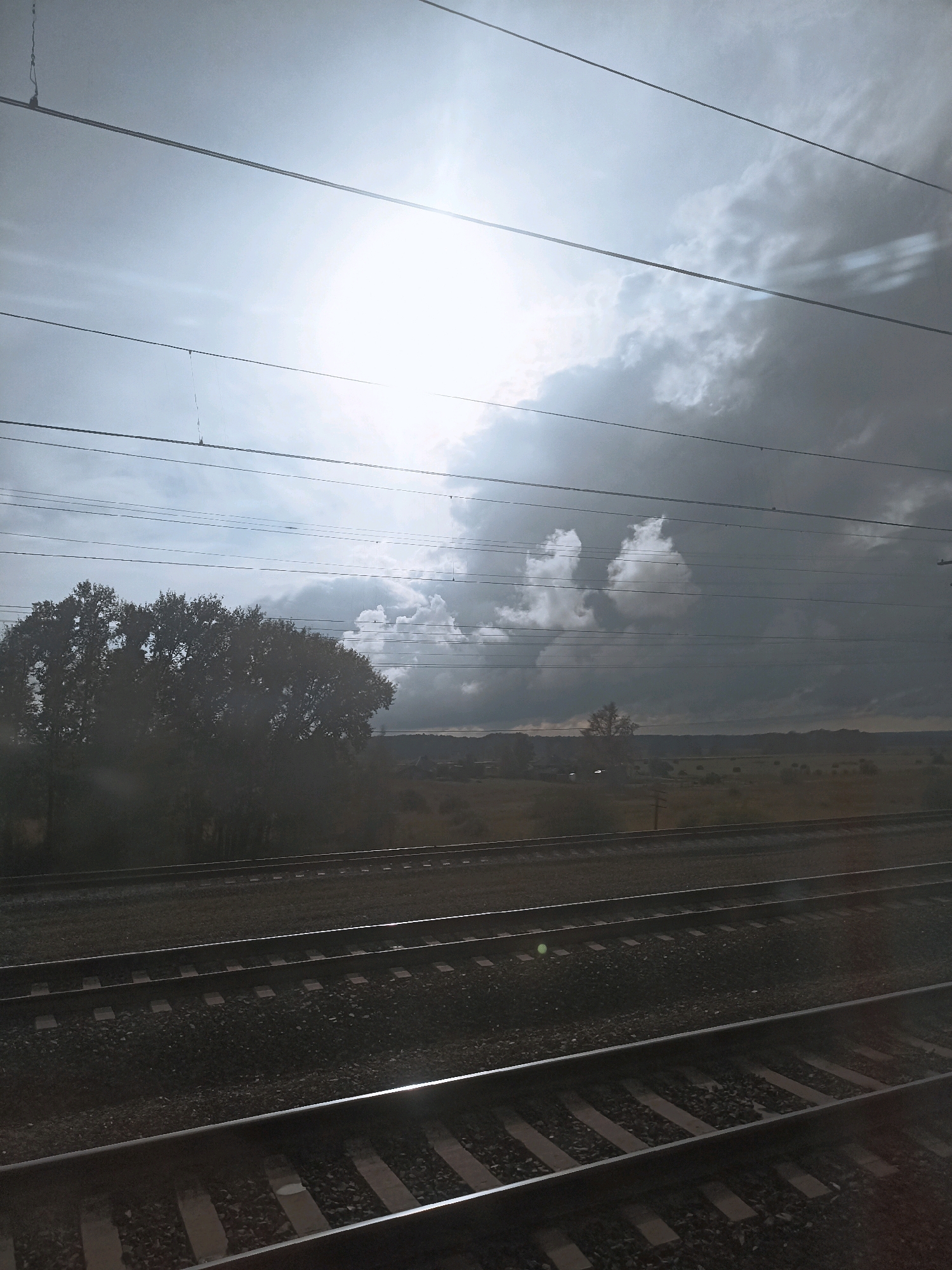 A view from a train window shows a cloudy sky with the sun peeking through. Below the sky are power lines, a line of trees, and fields of grass and dirt. In the foreground are train tracks. The scene is overcast, suggesting a cool day. The image appears to be taken in Kurundus, Russia.