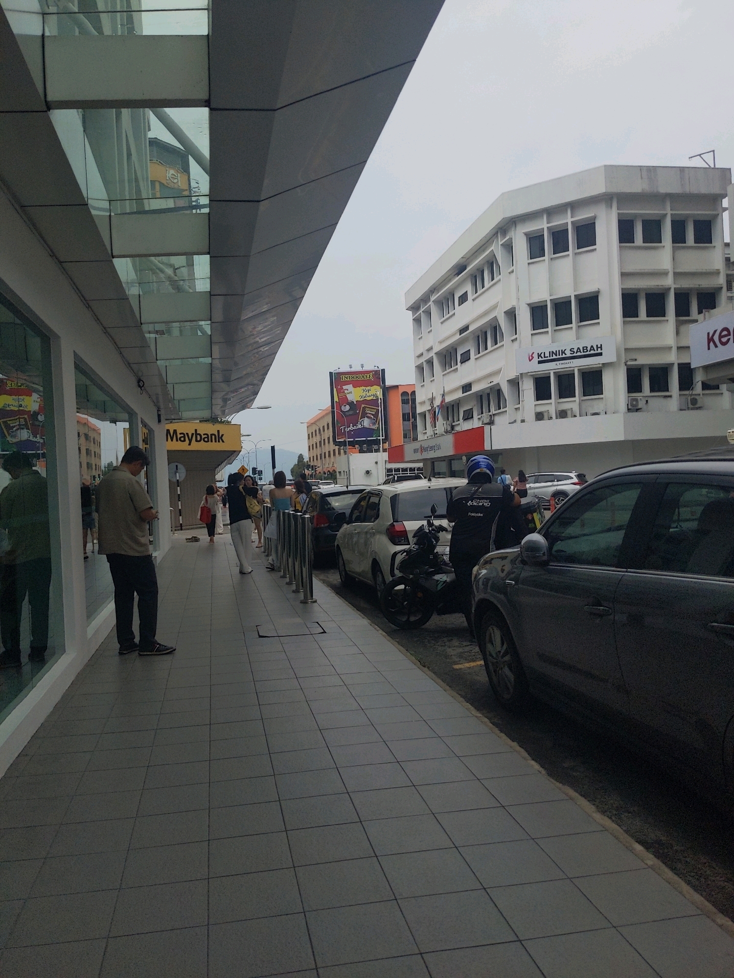The image depicts a street scene in Kota Kinabalu, Malaysia. A pedestrian sidewalk runs along the left side of the image, adjacent to a building with large glass windows. A yellow sign with the word "Maybank" is visible on this building. Several people are walking on the sidewalk, and one man in a light brown shirt is standing and looking at his phone.
Further down the street, another building with multiple windows and the text "KLINIK SABAH" is visible. Several cars are parked along the street, and a motorcycle is also present. The sky is overcast. A large billboard advertising "INDOCAFE" is situated to the right of the Maybank building. The scene appears to be an urban commercial area, with buildings, shops, and traffic.