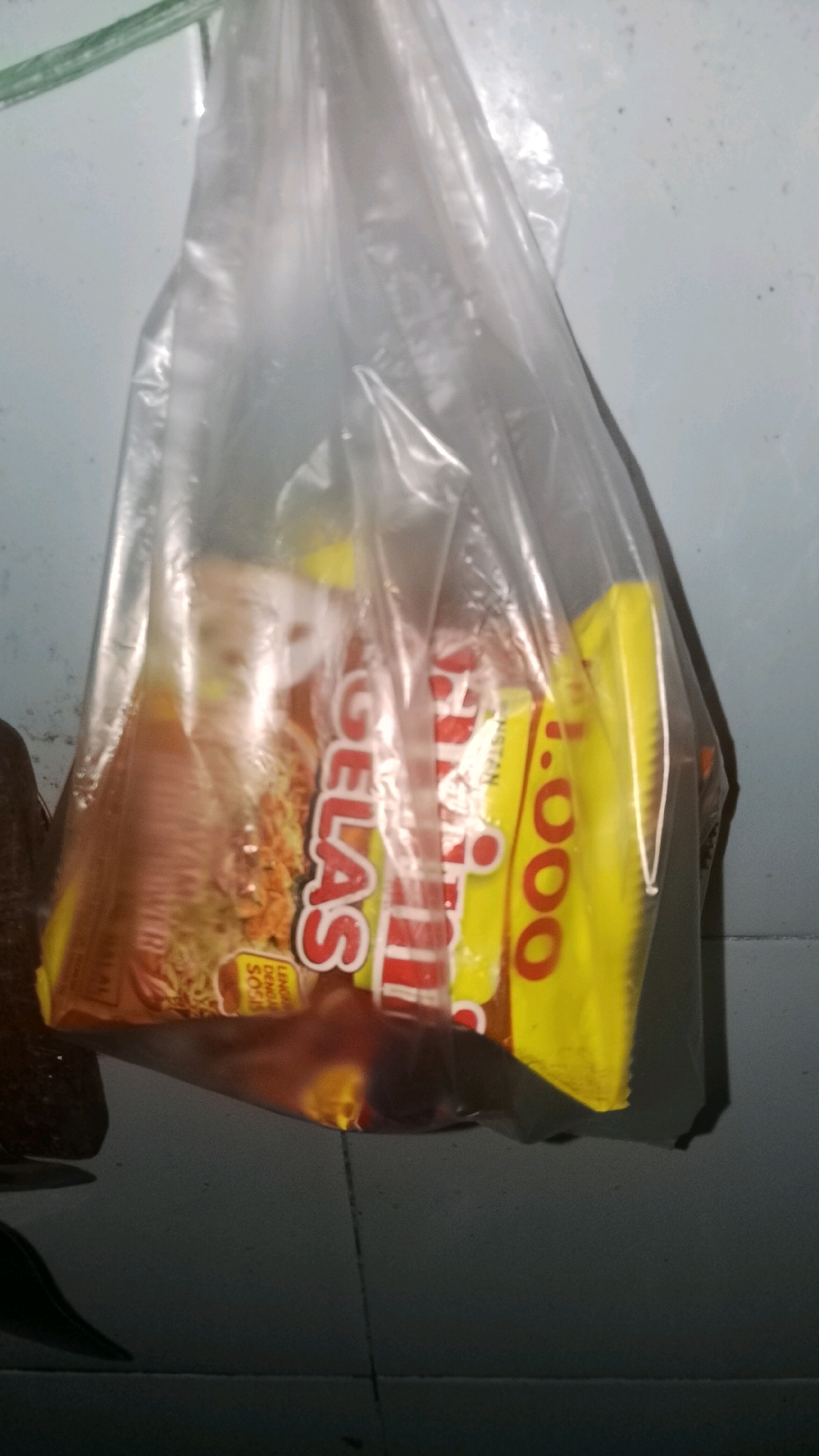 A plastic bag hangs from an unseen object, containing two packages of instant noodles. The packages are brightly colored, one with red and white lettering and the other yellow with red lettering. The bag is transparent and the contents are visible through it. The background is a light blue, tiled wall. The floor beneath the bag is dark and reflective, possibly a tiled surface. The image appears to have been taken in an indoor environment, likely a home or a shop, in the city of Mulyo Haji, Indonesia. There are no people or animals present in the image. The lighting is dim, and the image quality is low.