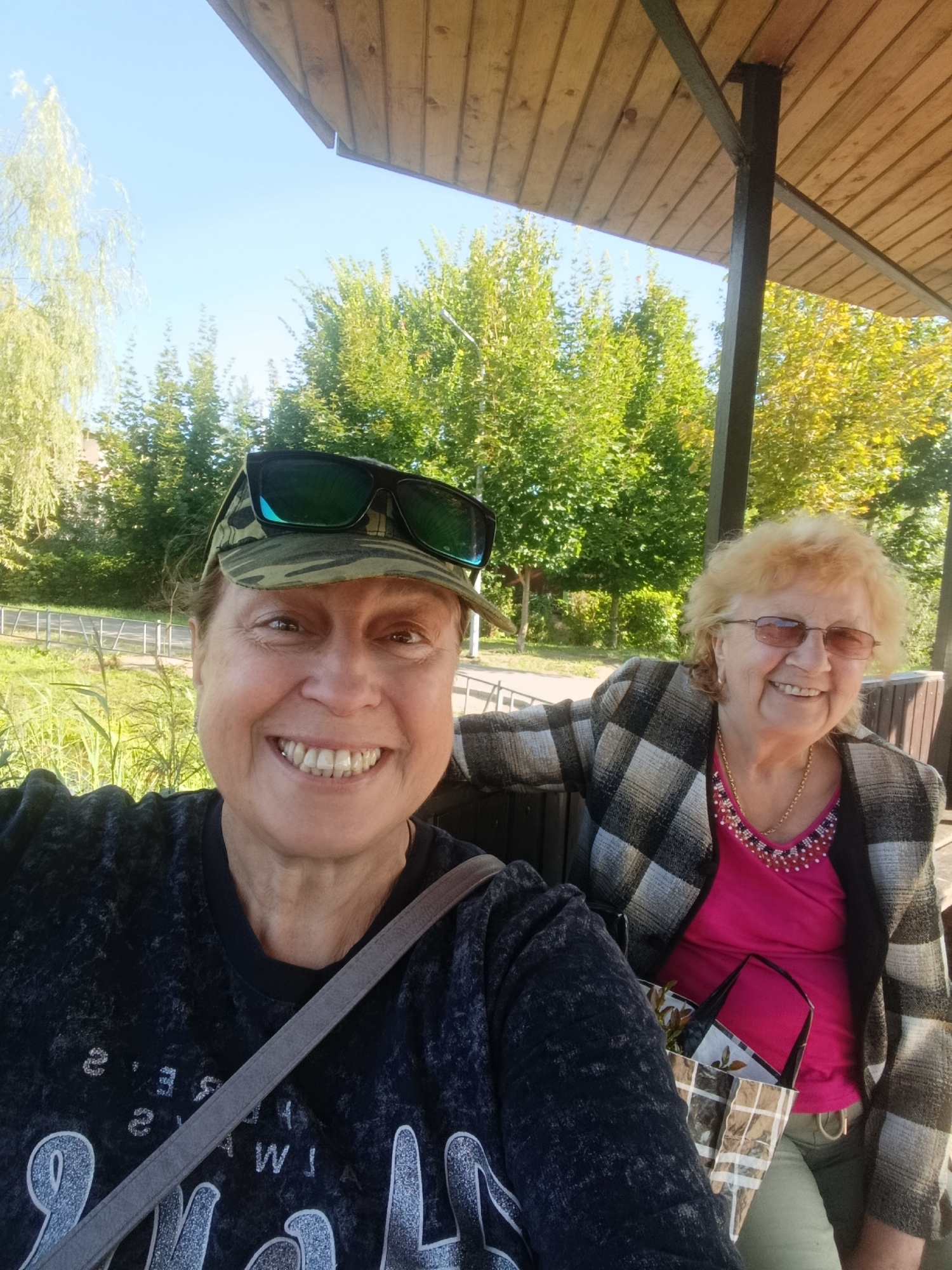 Two women are taking a selfie outdoors under a wooden roof. The woman in the foreground is wearing a camouflage baseball cap with sunglasses on top and a dark grey t-shirt. The woman behind her is wearing glasses, a pink top, and a checkered jacket. She is smiling and looking at the camera. The background shows trees and greenery, suggesting a park or natural setting. The image appears to have been taken during the daytime, with clear skies. The specific location is not identifiable as being in the city of Верея, Russia, based on the visual information provided.