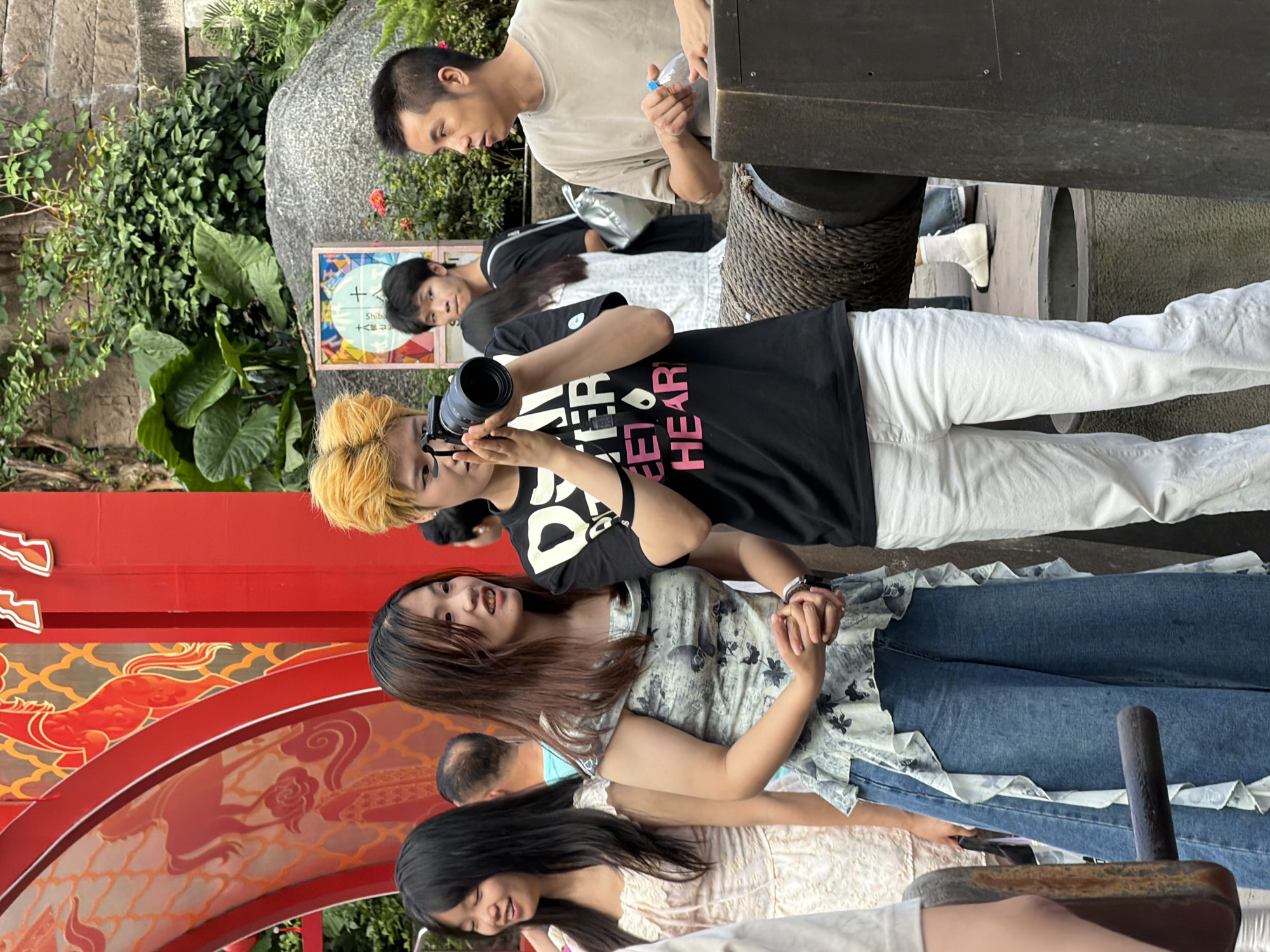 In Yuzhong District, China, a group of people are gathered outdoors near a red archway decorated with golden horse imagery. In the foreground, a young woman with dark hair, wearing a light-colored dress, smiles as she looks to her left. Next to her, a woman with long brown hair, dressed in a light blue floral patterned top and wide-leg jeans, clasps her hands together and looks towards the center. In the middle ground, a person with bright yellow hair, wearing a black t-shirt with pink text reading "SWEET HEART," holds a camera and appears to be taking a photo. Behind them, other individuals are visible, including a young man in a black t-shirt and a man in a beige t-shirt holding a water bottle. The background features lush green foliage, a large rock formation, and a stone wall. The overall scene suggests a public gathering or event.