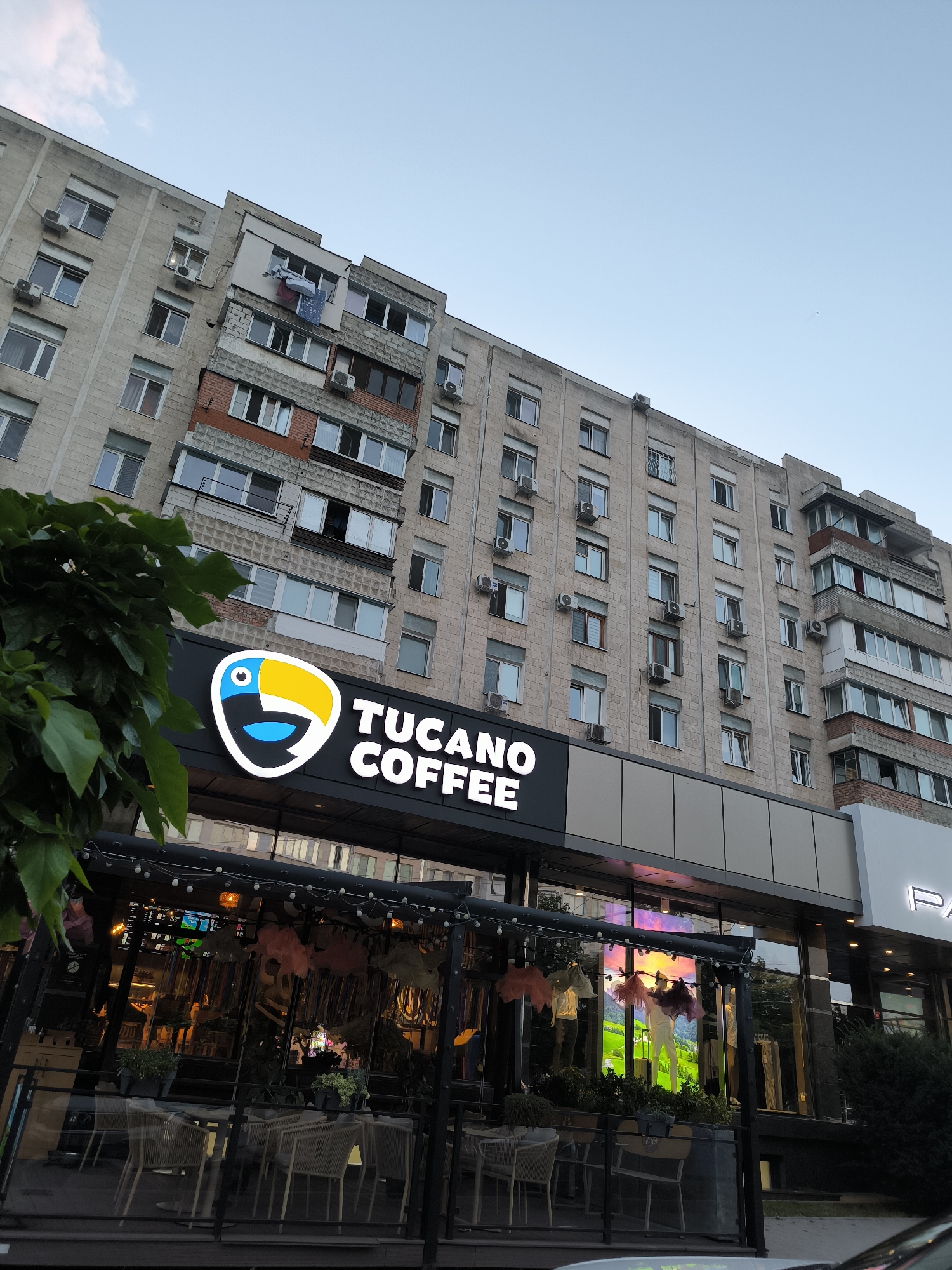 The image shows the exterior of a "Tucano Coffee" establishment located in Chișinău, Moldova. The coffee shop is situated at the ground level of a multi-story apartment building, characterized by its light-colored, brick-like facade and numerous windows, many of which are equipped with air conditioning units. The coffee shop features a prominent sign with the "Tucano Coffee" logo, which includes a stylized toucan. In front of the coffee shop, there is an outdoor seating area with tables and chairs, partially enclosed by glass. The overall scene depicts a typical urban environment with residential buildings and commercial establishments.