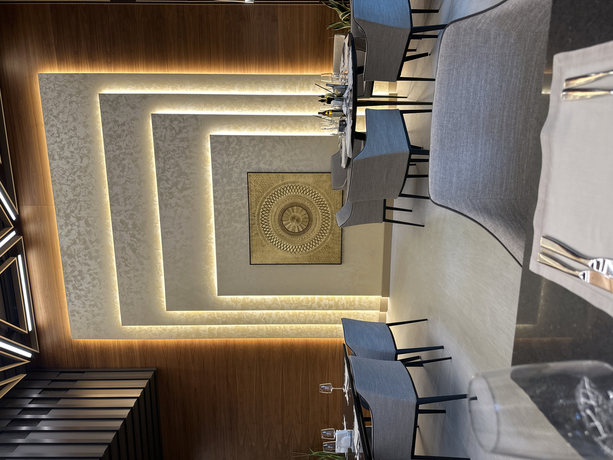 This media file captures the elegant interior of a modern restaurant or dining area, presented in a portrait orientation. The scene is devoid of people, suggesting a moment before service or during off-hours.
The most striking feature is the back wall, which is adorned with light-colored, textured panels arranged in a geometric, layered pattern of concentric rectangles. Warm, indirect LED lighting emanates from behind these panels, creating a soft, inviting glow and highlighting the wall's texture. Centered on this illuminated feature is a framed, square artwork displaying an intricate, circular, golden-colored design, resembling a mandala or sunburst pattern.
To the left, a section of the wall is paneled with warm-toned wood, adding a natural and sophisticated element to the decor. Further to the left, another modern architectural element with linear white lighting is partially visible.
The dining area is furnished with several tables featuring reflective black surfaces, surrounded by grey upholstered chairs with sleek black metal frames. Many tables are meticulously set with clear wine glasses, water glasses, white napkins, and silver cutlery (forks and knives), indicating readiness for dining. A green potted plant adds a touch of natural life to the setting.
The lighting throughout is artificial, predominantly warm and indirect, contributing to an upscale and tranquil ambiance, typical of an evening dining setting. The overall impression is one of contemporary design and refined taste, fitting for a modern establishment in a location like Kazanlak, Bulgaria. No visible text is present in the image.