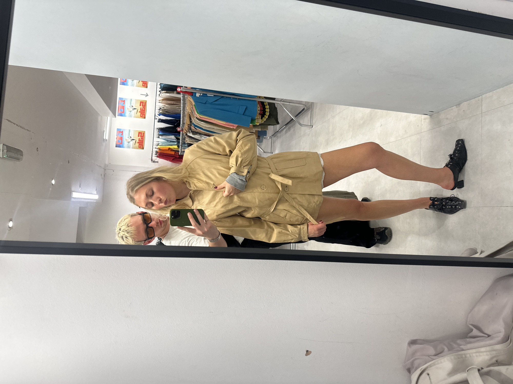 In a brightly lit retail store, likely a vintage clothing shop in London, United Kingdom, a young woman with blonde hair is posing for a selfie in a mirror. She is wearing a tan trench coat over white shorts and black heeled mules. A man with bleached blonde hair and sunglasses stands behind her, also reflected in the mirror. The store's interior is visible, with racks of clothing and posters on the wall, suggesting a bustling marketplace.