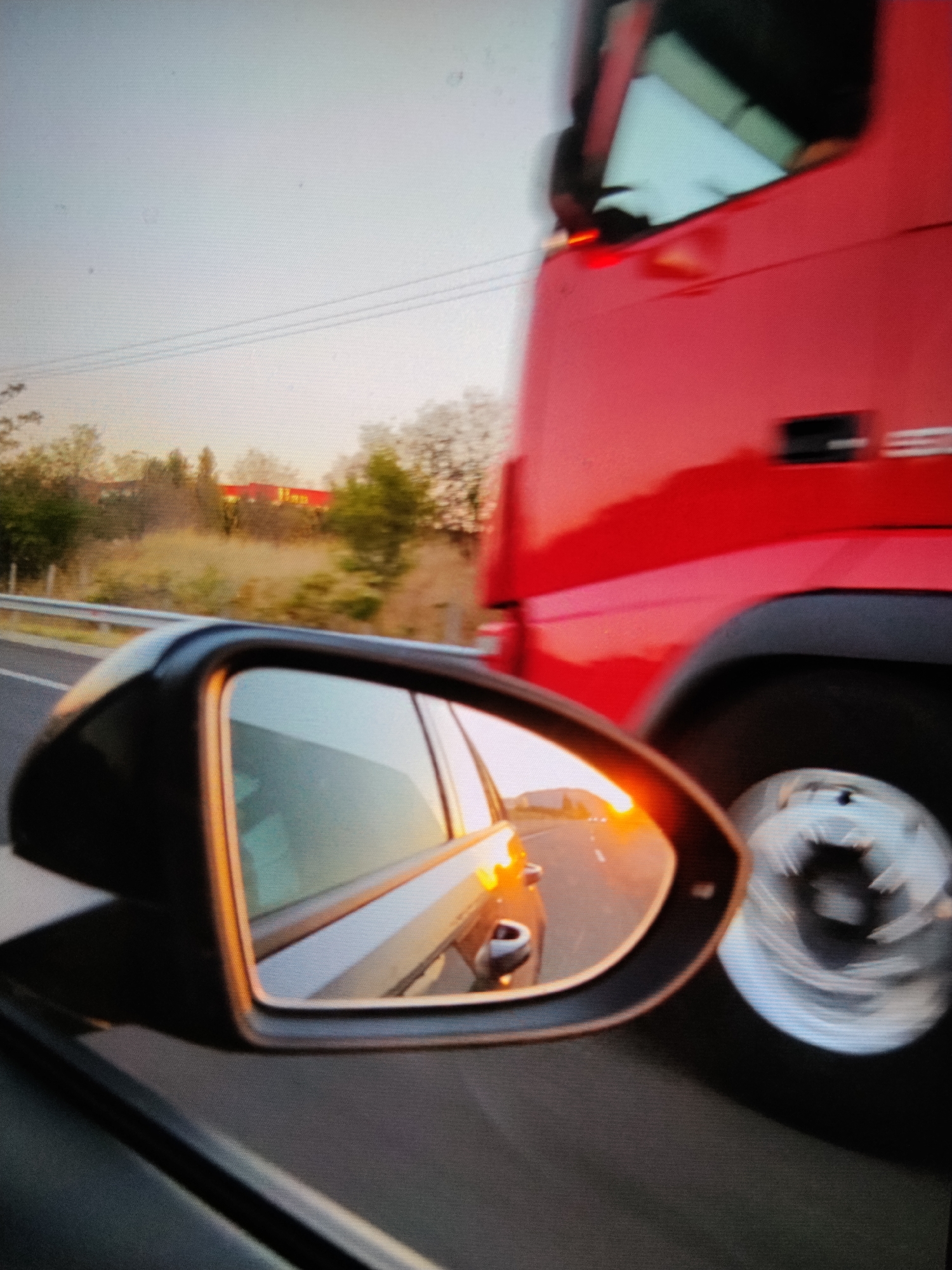 The image shows a red truck passing by a car, as seen in the car's side mirror. The mirror reflects the car's side and a road with an orange glow, likely from the setting sun. The truck is large and dominates the right side of the frame, with a visible wheel. Outside the car window and further back in the image, there's a glimpse of a highway with a railing, some trees, and a building with a distinct red roof. Based on the specific red roof and surrounding environment, the location is on the highway towards Athens, Greece, and the red roof building appears to be part of the McArthurGlen Athens outlet complex.