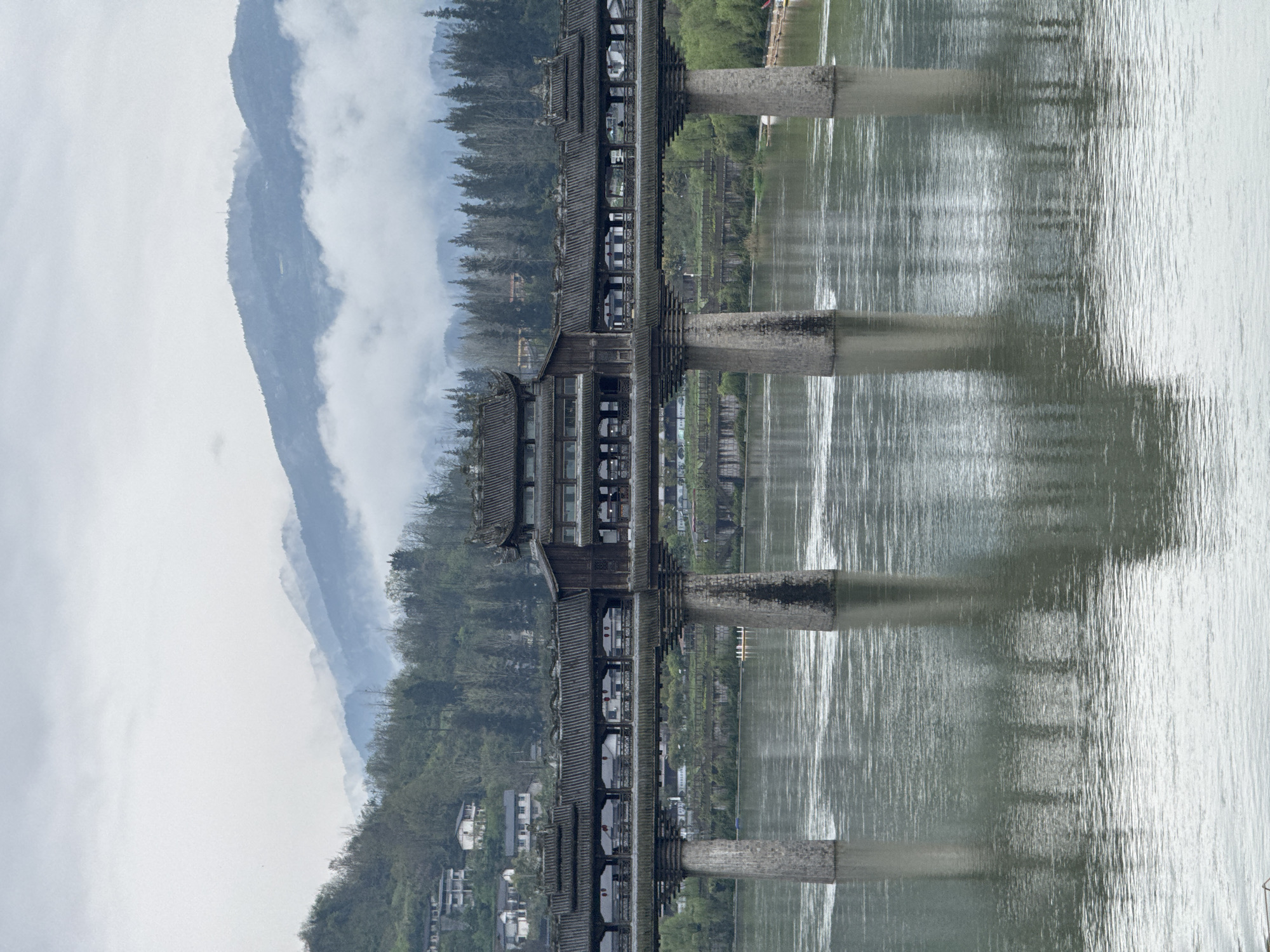 The image shows an ancient covered bridge spanning a river in Zhuoshui, China. The bridge features traditional Chinese architecture, with a dark-tiled roof and wooden structure. The river reflects the bridge and surrounding scenery, creating a symmetrical composition. Mountains, partially obscured by clouds, rise in the background. Buildings are visible on the riverbank near the base of the mountains. The sky is overcast, contributing to the subdued lighting of the scene.