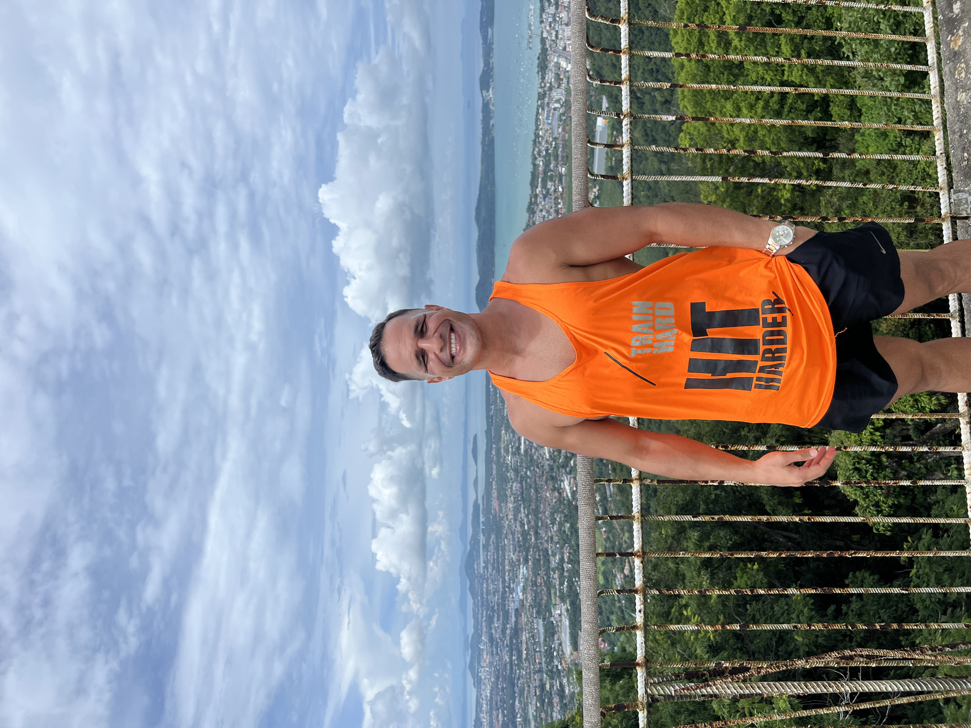 A man stands on a railing overlooking a city and ocean, with a cloudy sky. He is wearing an orange tank top that says "TRAIN HARD HIT HARDER" and black shorts. He has a watch on his left wrist and his right hand is in his pocket. The city is visible in the background, with buildings and a bay with boats. The man is smiling. The location is likely Karon Viewpoint, Karon, Thailand.