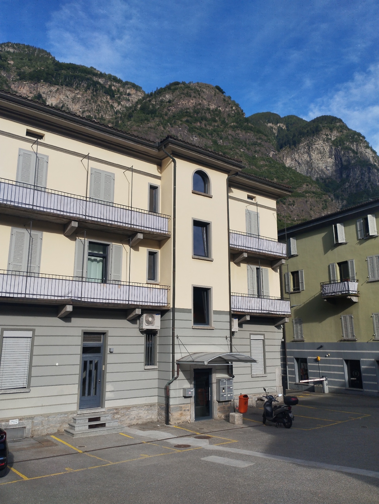 The image depicts a street scene in Biasca, Switzerland, under a clear blue sky with scattered clouds. In the foreground, a paved area with yellow parking lines is visible, with a parked black scooter on the right side. Two buildings are the main focus. The building on the left is a multi-story structure with a cream-colored facade and grey horizontal siding on the lower levels. It features multiple balconies with grey railings and white coverings, and numerous windows with grey shutters. An outdoor air conditioning unit is mounted on the side of this building. To the right of the cream-colored building is a second, taller building with a muted green facade, also with multiple windows and balconies. In the background, a steep, rocky mountain covered in dense green vegetation rises prominently, dominating the upper portion of the image. The overall scene suggests a residential or commercial area situated at the base of a mountain.