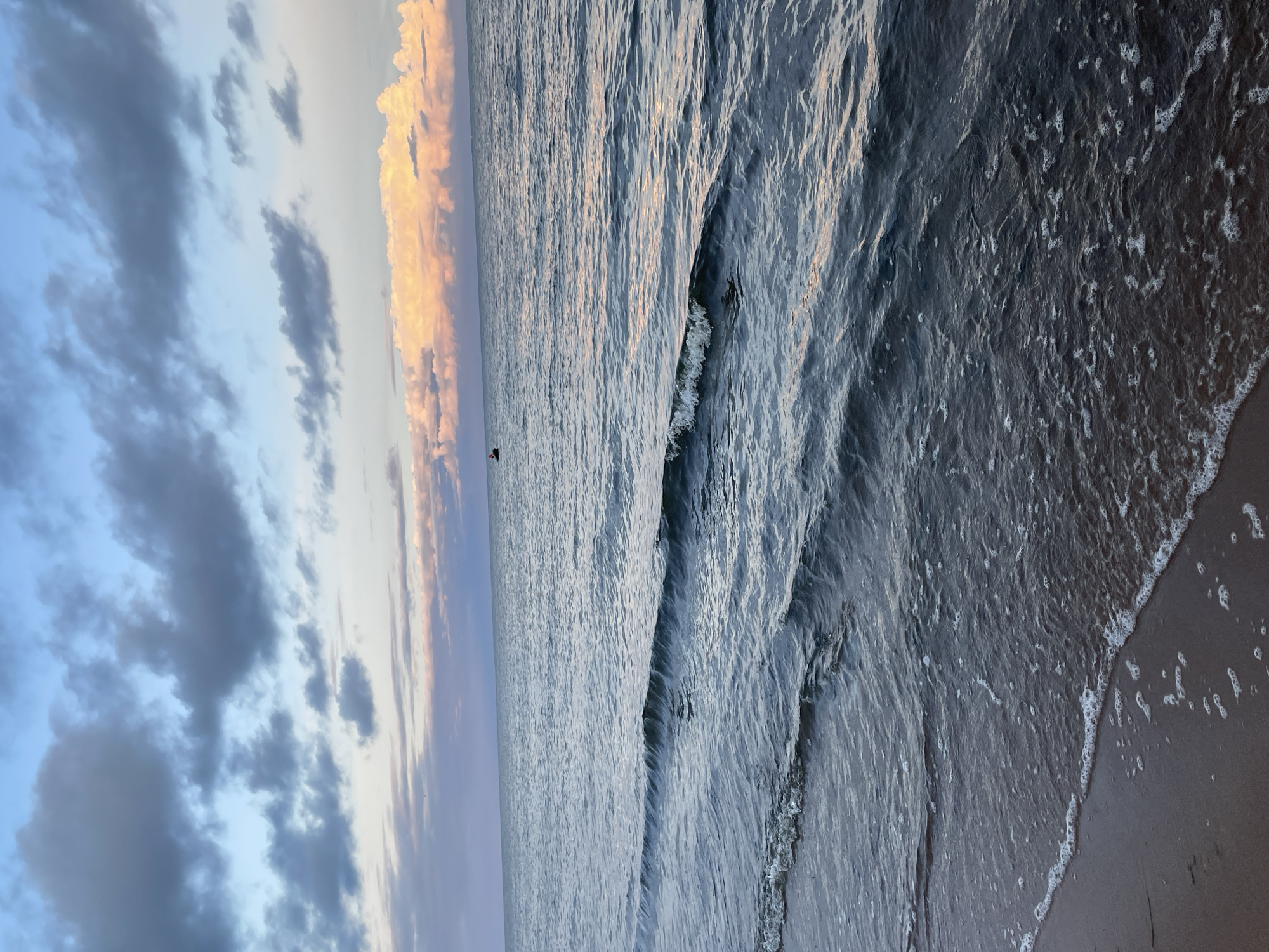 The image depicts a serene seascape at dusk, likely near Zelenaya Roshcha, Russia. The sky is a canvas of soft blues and grays, with streaks of pale pink and orange from the setting sun illuminating the distant clouds. A few dark, wispy clouds are scattered across the upper portion of the frame. The sea is calm, with gentle waves lapping at the sandy shore in the foreground. The water is a deep, muted blue, reflecting the colors of the sky. A small boat is visible in the distance, appearing as a dark silhouette against the horizon. The beach itself is sandy, with the wet sand at the edge of the water showing faint patterns from the receding waves. The overall atmosphere is tranquil and peaceful, capturing the quiet beauty of the coastline as day transitions into night.