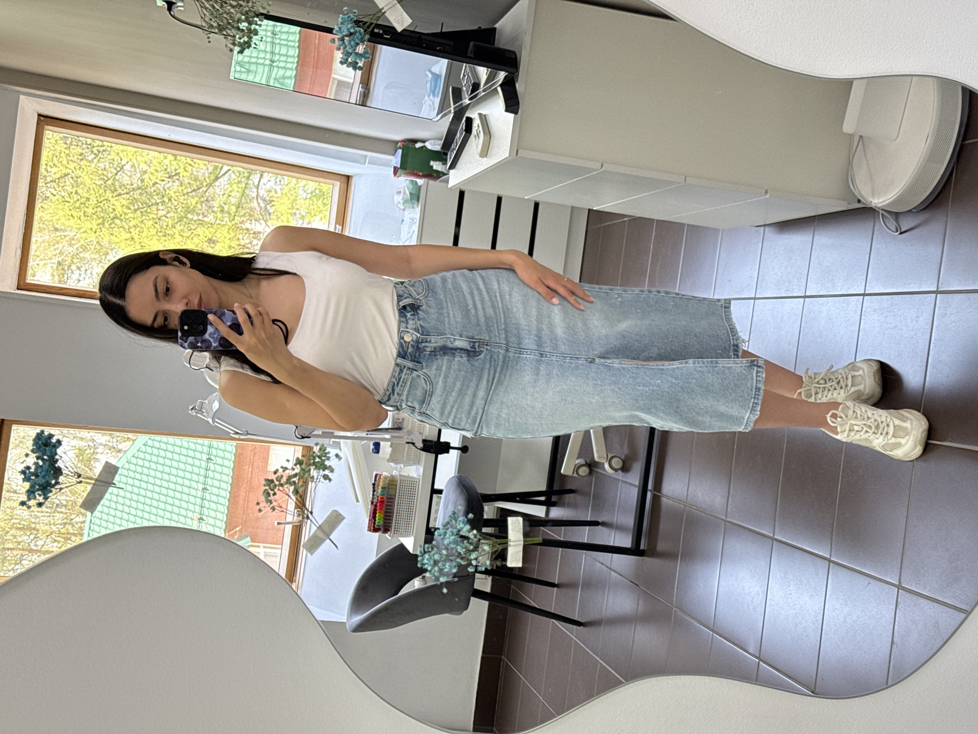 A young woman is taking a selfie in a mirror in an interior space. She is wearing a white tank top, a light blue denim skirt, and beige sneakers. She is holding a smartphone with a dark patterned case. Reflected in the mirror is a room with brown tiled floors, a grey chair with blue flowers on it, a white desk with various items, including a computer monitor, and two windows. Outside the windows, green trees and buildings can be seen. Based on the presence of buildings and trees, it is impossible to say whether or not this is Chaikovsky, Russia.