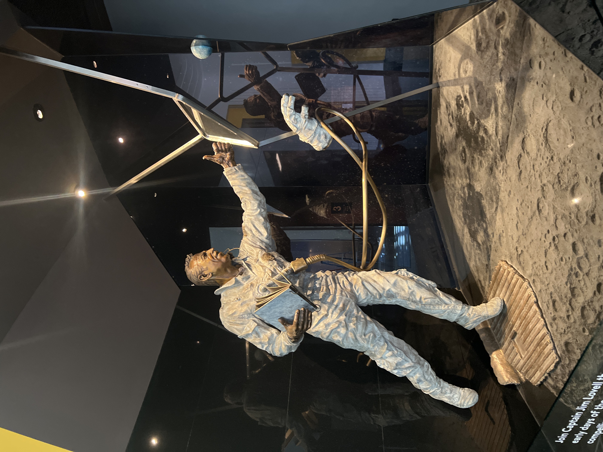 The image shows a museum exhibit featuring a statue of Captain Jim Lovell, a NASA astronaut known for his role in the Apollo 13 mission. The exhibit is likely located in Chicago, as the text visible at the bottom mentions Captain Jim Lovell and his compelling story. The statue depicts Lovell in a spacesuit, holding a book or manual, with one arm raised towards a globe. The figure stands on a textured base resembling the lunar surface. The statue and surrounding elements are enclosed in a glass case with black panels, creating reflections. The exhibit also includes a metallic framework with a small blue sphere.