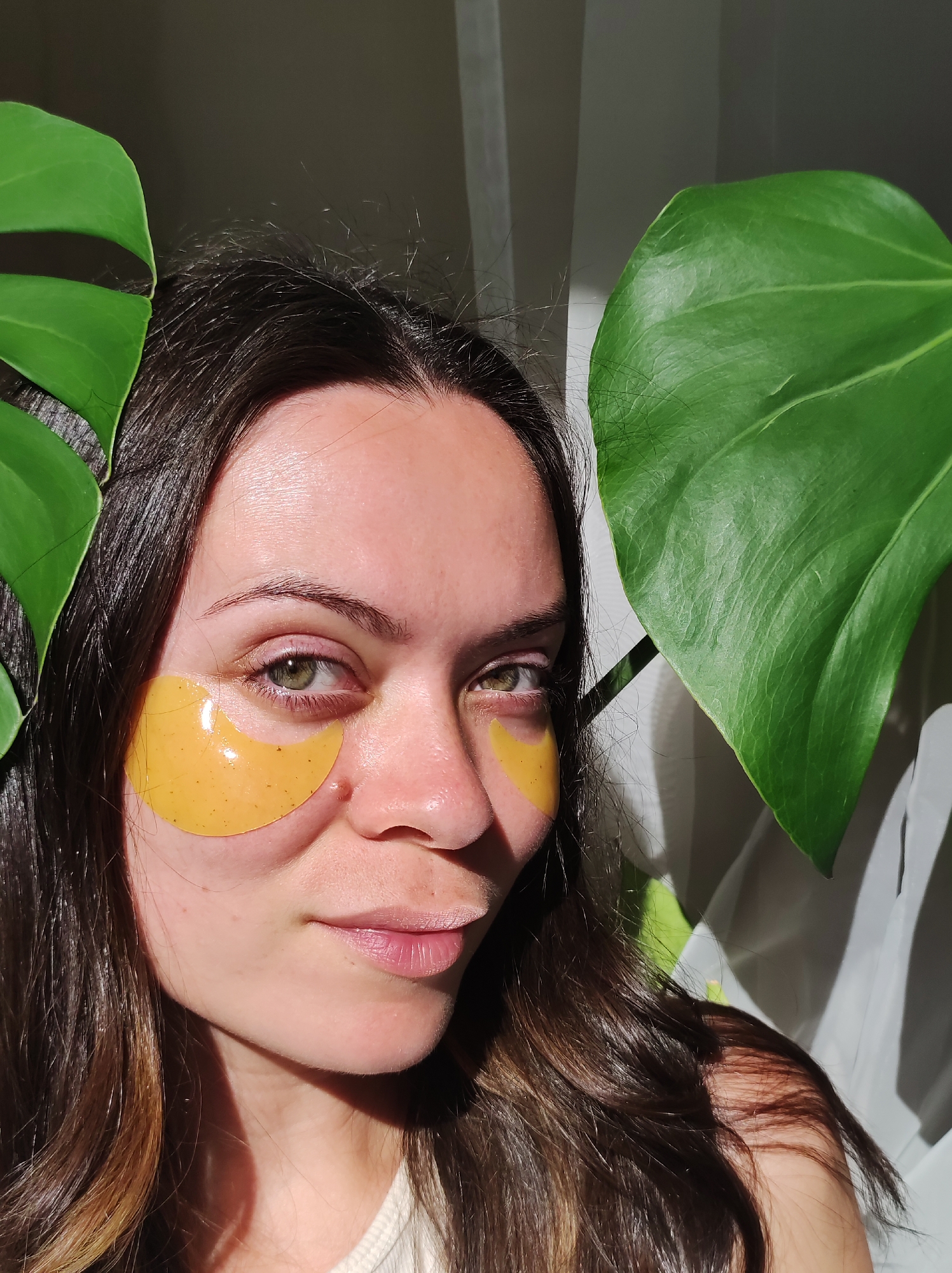 A close-up, head-and-shoulders portrait shows a young woman with brown hair and green eyes, wearing yellow under-eye patches. She is positioned slightly to the left of center, with large green leaves from a Monstera plant framing her face on either side. Sunlight casts shadows across her face and hair. The background is out of focus, appearing to be an indoor setting with draped white fabric and a dark wall. There is no indication of the city of Chaikovsky, Russia, within the image.