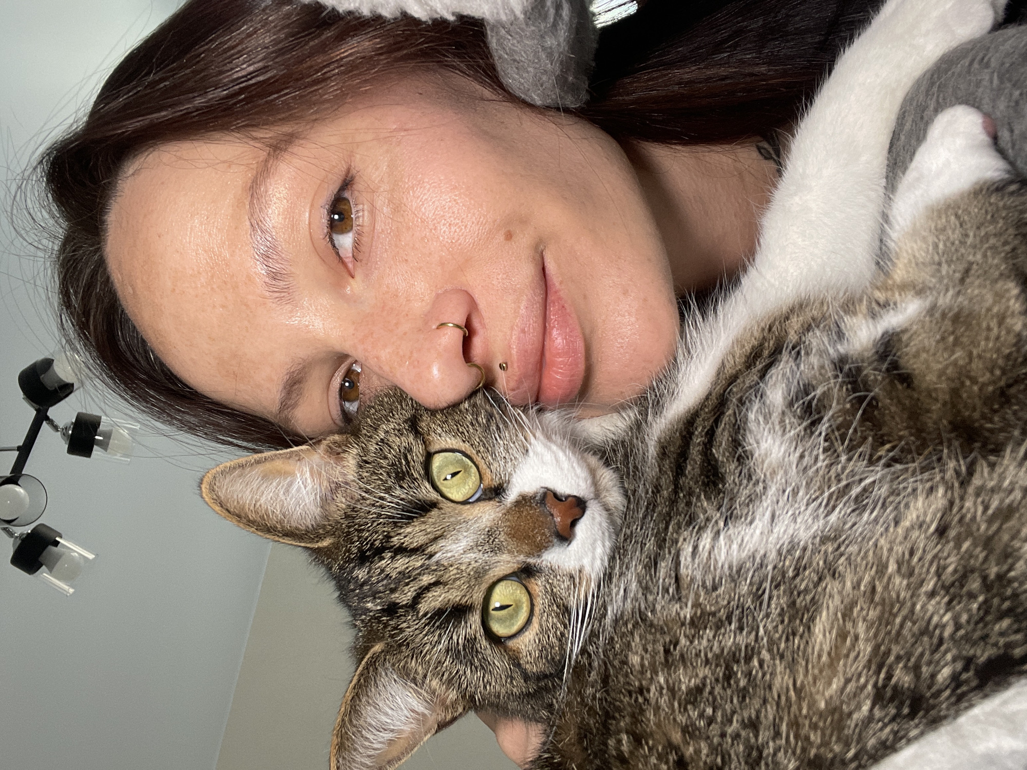 A woman with brown hair is holding a tabby cat close to her face. She has fair skin, freckles, and two gold nose piercings. The cat has green eyes and is looking directly at the camera. The background is a mix of gray fabric and a blurry room with a ceiling light fixture. The scene appears to be indoors. The location is not discernible as Izhevsk, Russia from the image alone.