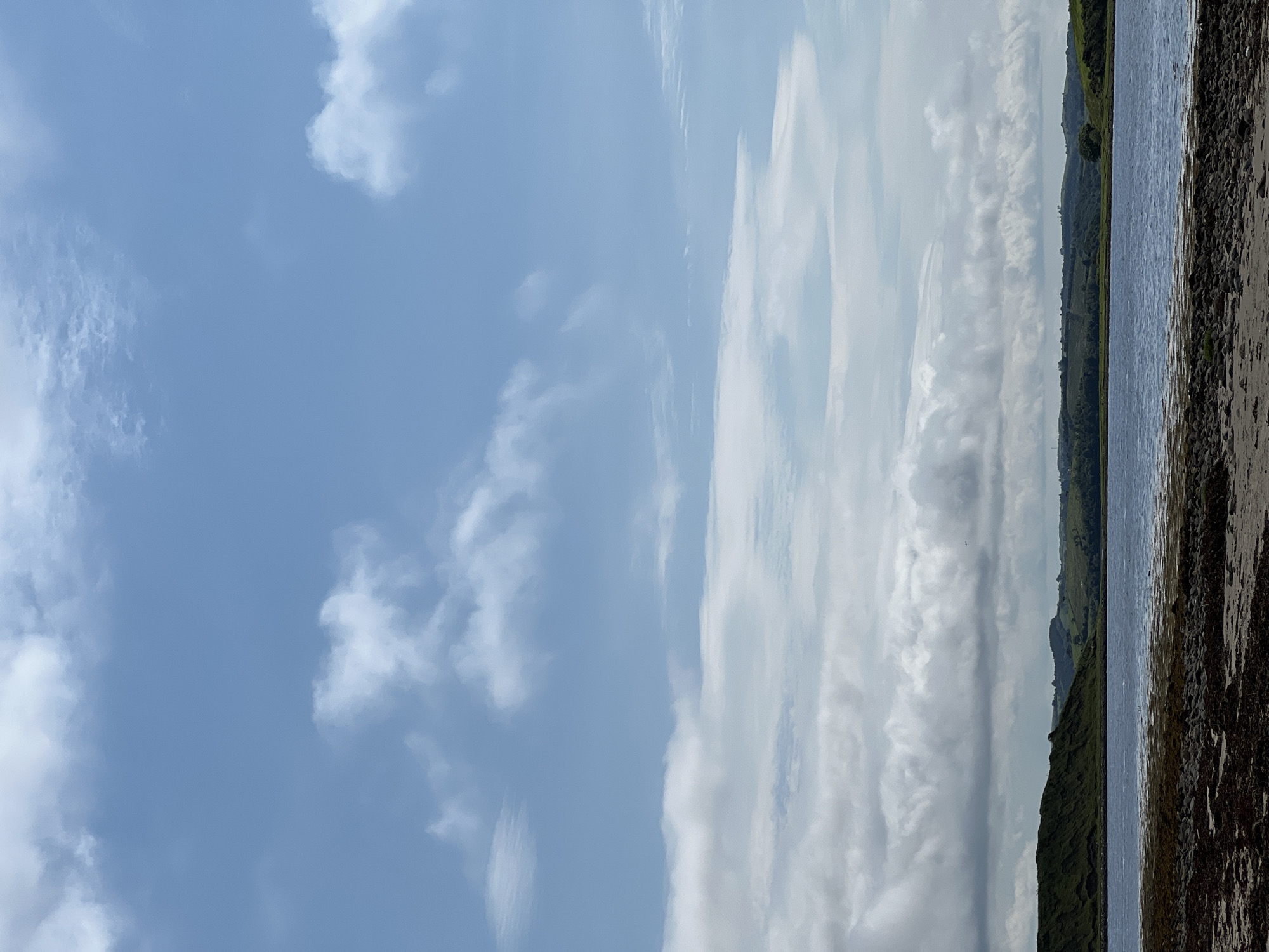 The image depicts a serene coastal landscape. The foreground features a shoreline composed of dark rocks and seaweed, transitioning into a body of water that appears to be a loch or bay. In the mid-ground, rolling green hills form the coastline, with some appearing to be covered in heather. The sky above is a pale blue, dotted with various types of clouds, some wispy and white, others more substantial and grey. The overall impression is one of natural beauty and tranquility. The location is identified as Dunuisce, Ireland. There are no people or animals visible in the image, and no discernible actions or interactions are taking place.