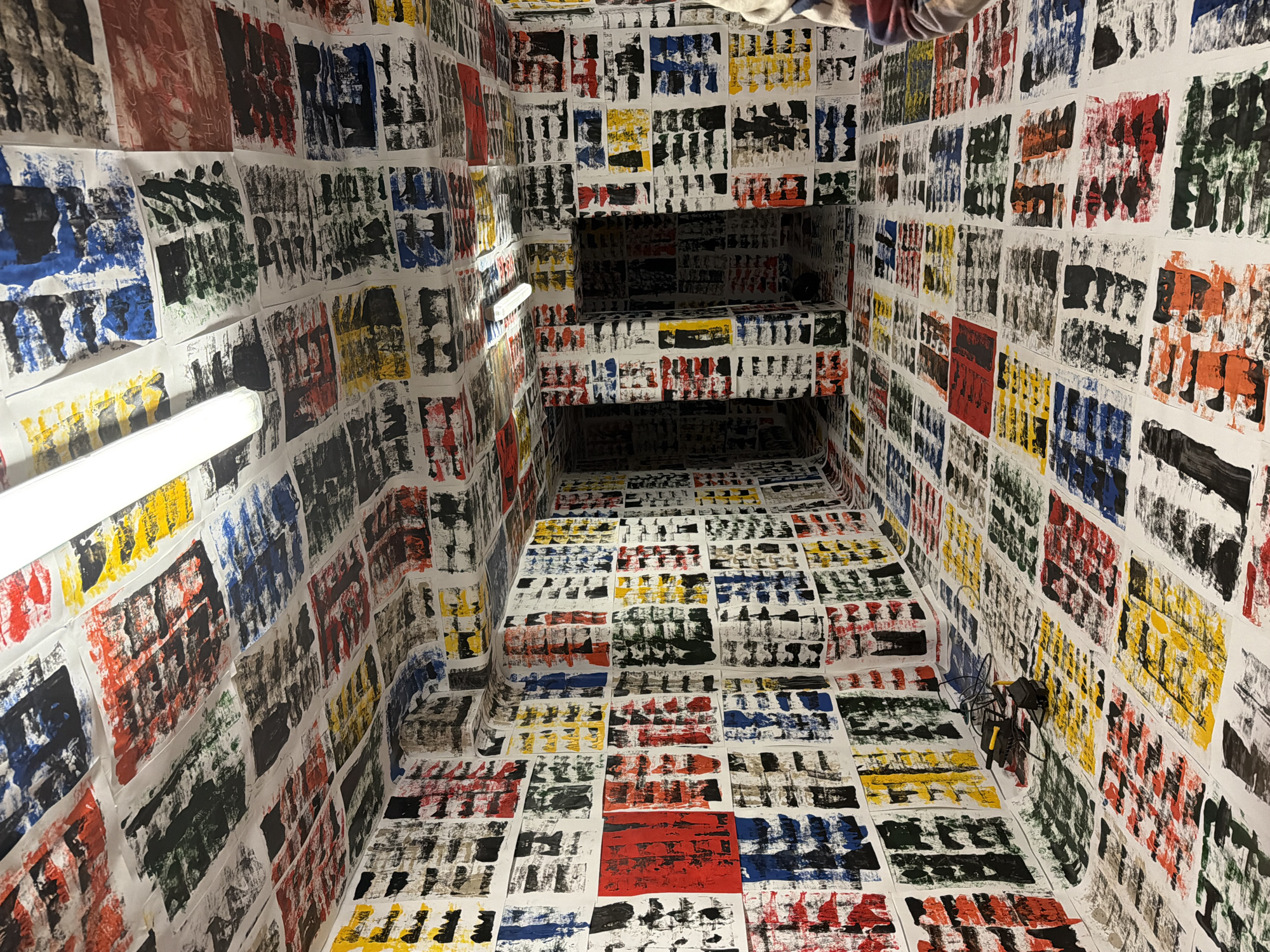 The image shows an interior space completely covered with white sheets of paper. Each sheet contains abstract block prints in various colors including black, red, blue, green, and yellow. The space appears to be a narrow passage or small room with a stepped recess or opening further in. Two fluorescent tube lights are affixed to the walls. A small, dark object, possibly a bicycle, is visible on the wall on the right side. The location is not discernible from the image.