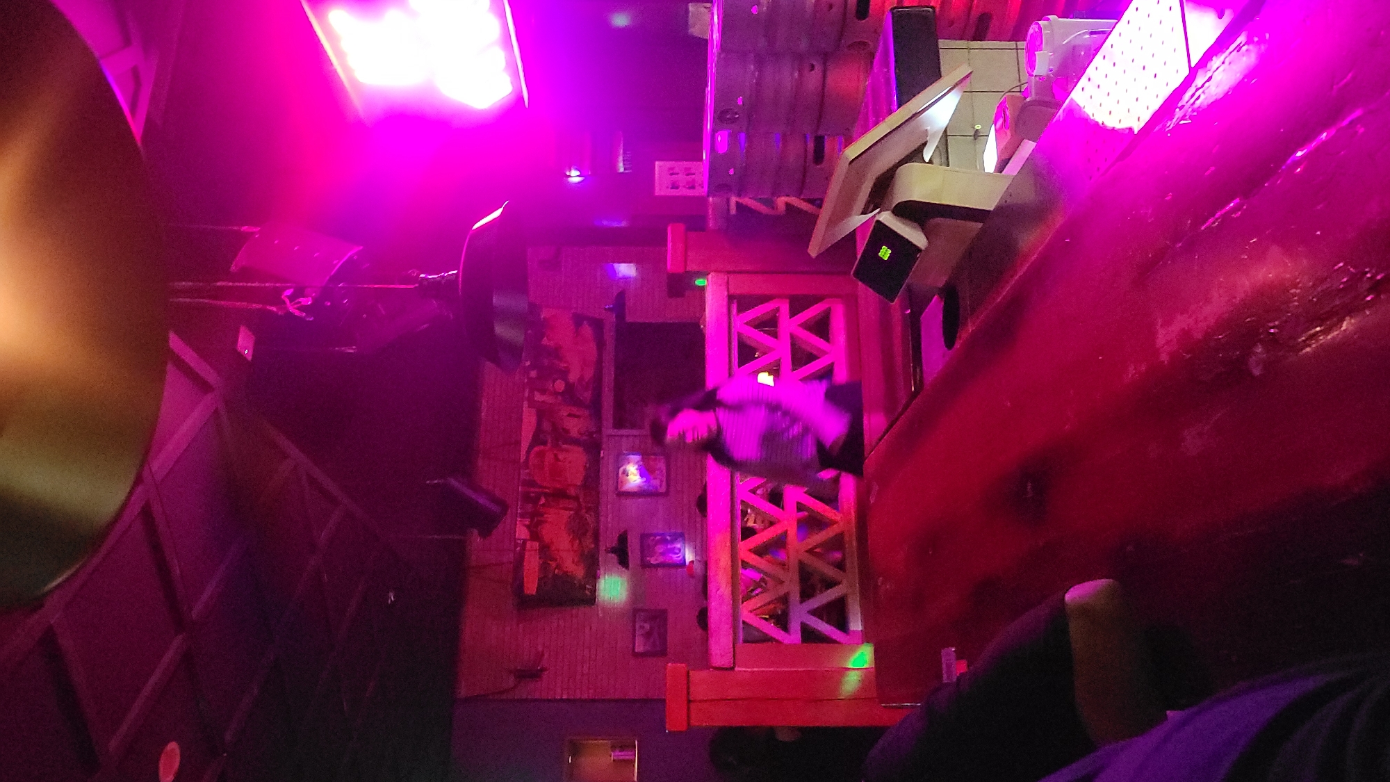 This is an interior shot of a dimly lit entertainment venue, saturated with vibrant magenta and pink lighting, suggesting a nightclub, bar, or themed attraction. The image appears to be rotated 90 degrees clockwise.
In the immediate foreground, to the right, a large, curving, reddish-brown structure dominates the frame, possibly part of a booth, wall, or a large piece of equipment. Its surface reflects the intense pink light, showing some texture or wear. Part of a person's shoulder and arm, clad in dark clothing, is visible at the very bottom right, blending into the shadows.
Further into the scene, several bright magenta light fixtures, possibly LED panels, emit a strong glow from the upper left and upper right sections of the image. On the left side, a section of a wall with what appears to be a textured or paneled surface is visible, adorned with multiple framed pictures or artworks of varying sizes.
In the mid-ground, a person is seen walking or standing near a distinctive wooden partition or doorway with a geometric, criss-cross (X-shaped) pattern. This person is wearing a lighter-colored top and dark pants, and their figure is illuminated by the pink light. Several small, bright green lights are scattered throughout the scene, possibly indicating sensors, indicators, or decorative elements. On the right, near the large reddish structure, a small, light-colored device with a green light is mounted. In the upper-mid-ground, multiple monitors or screens with white light are visible.
The overall atmosphere is energetic and dynamic, defined by the striking color palette of the lighting. No discernible text is visible within the image.