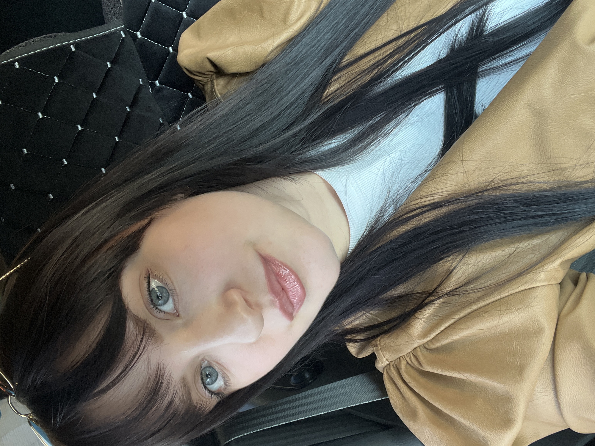 The image displays a young woman with long, straight dark hair, blue-grey eyes, and fair skin, looking directly at the camera with a neutral expression. Her head is tilted, suggesting she is leaning back or lying down. She is wearing a white top and a tan or camel-colored jacket, which appears to be made of faux leather and features gathered details on the sleeve. Her lips are accentuated with a glossy, pinkish-nude lipstick.
The setting is the interior of a vehicle, likely a car. Background elements visible include black quilted material, which could be floor mats or seat covers, and a dark grey seatbelt diagonally crossing the lower right portion of the frame. The lighting suggests it is daytime. The scene is located in Chaikovsky, Russia. There is no visible text in the image.