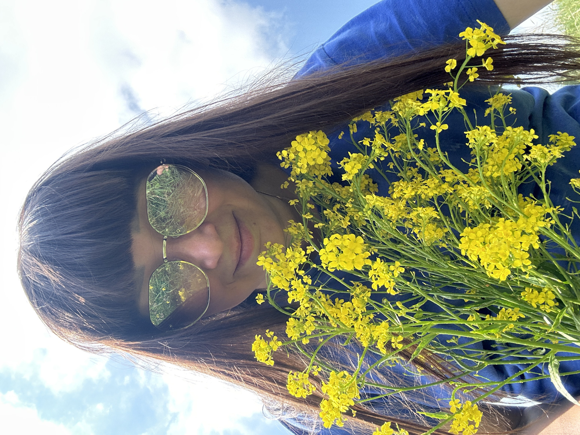 The image is a close-up, low-angle shot of a woman wearing sunglasses and a blue t-shirt, holding a bunch of small yellow flowers. Her dark brown hair frames her face, and her bangs are visible. The sunglasses reflect a sunny sky with clouds and green vegetation. The background is a clear blue sky with some scattered white clouds. The image appears to be taken outdoors, possibly in a field or park, given the presence of flowers and the natural lighting. The location is identified as Buranovo, Russia, though no specific city landmarks or environmental features confirm this. The focus is on the woman and the flowers, with the sky as the backdrop.