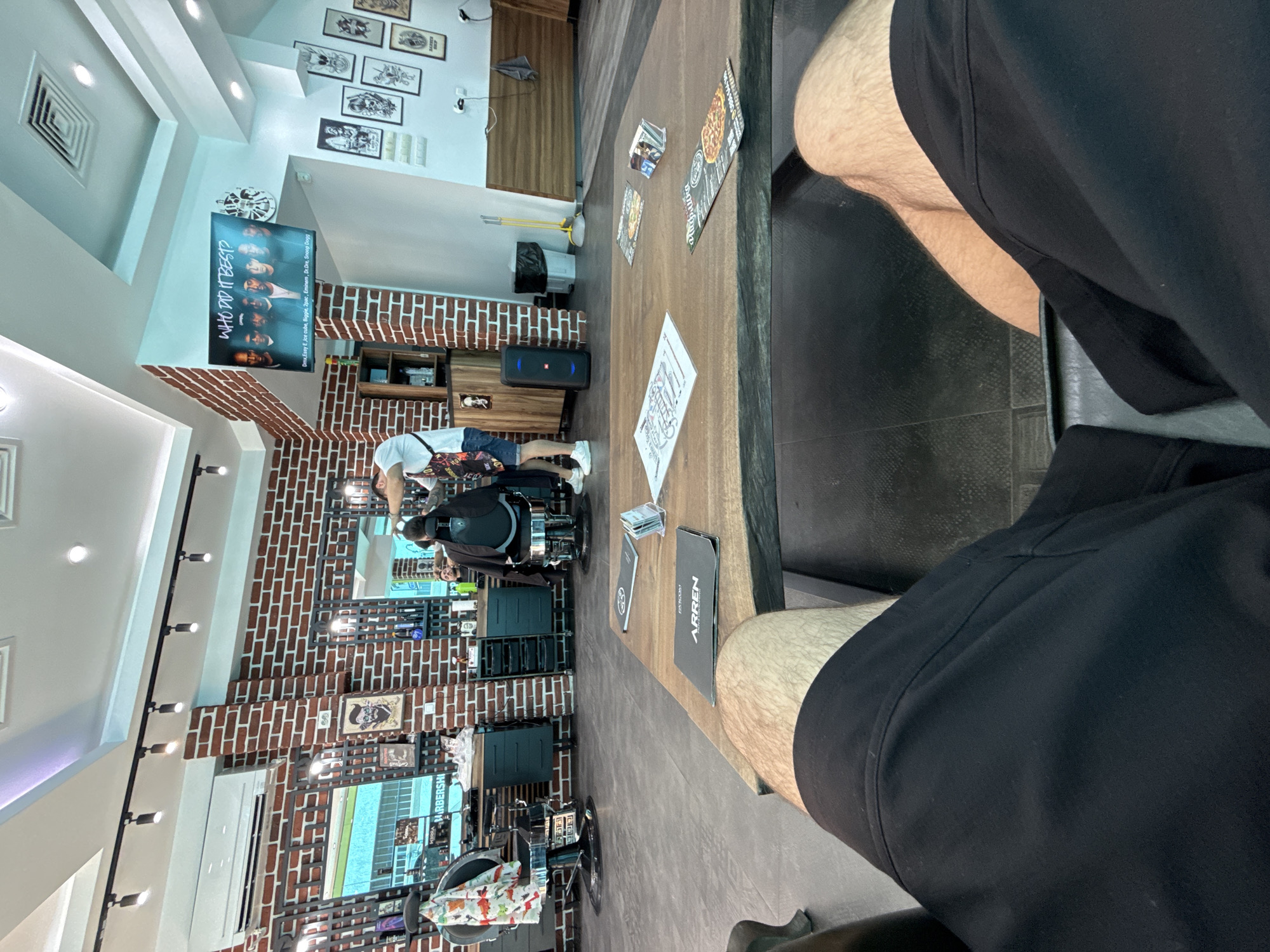 The image captures the interior of a modern barbershop, likely in the city of Shumen, Bulgaria, as indicated by the context provided. The scene is viewed from the perspective of someone seated at a large, rustic wooden table in the foreground, with their legs and black shorts visible. On the table are various items including menus, flyers, and a small holder with cards.
The barbershop itself features exposed brick walls, a common aesthetic in such establishments. Multiple barber chairs with chrome accents are visible, some with colorful capes draped over them. A barber is actively attending to a customer seated in a chair, indicating an ongoing service. A large screen mounted on the wall displays an image with text that reads "WHO DID IT BEST?" alongside portraits of several individuals, possibly musicians or artists. Decorative posters and artwork adorn the walls.
The overall environment suggests a busy and well-equipped barbershop, with elements of contemporary design blended with industrial touches. Lighting appears to be a mix of overhead fixtures and track lighting. The perspective implies the photographer is waiting or observing the services being rendered.