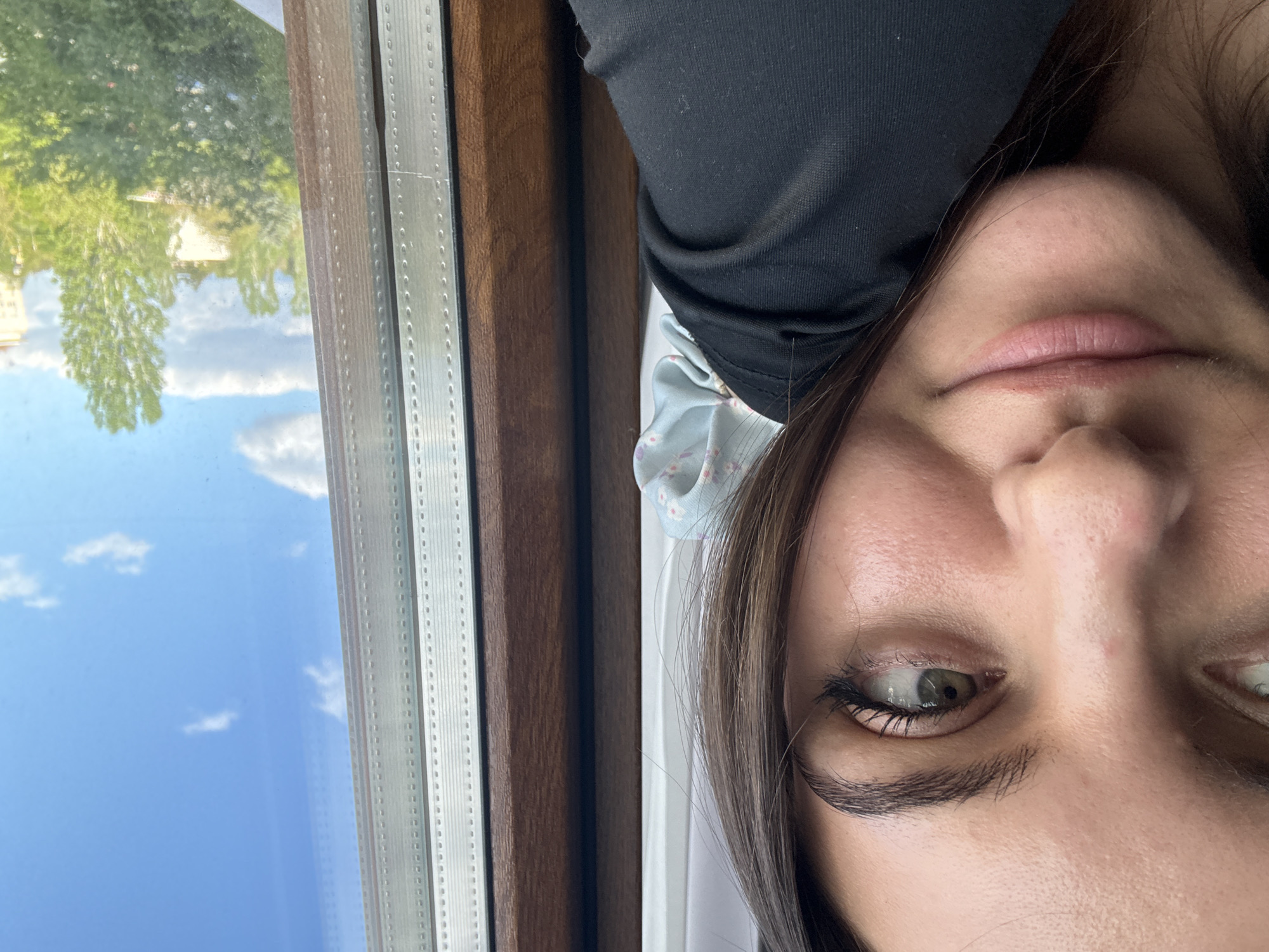 A close-up portrait shows a woman indoors, possibly near a window. Part of the window is visible, showing a reflection of trees and a blue sky with clouds. The woman has brown hair and light skin. She is wearing a dark shirt and a light blue hair accessory. The location is unknown, and there is no information to suggest that the photo was taken in Sofia, Bulgaria.