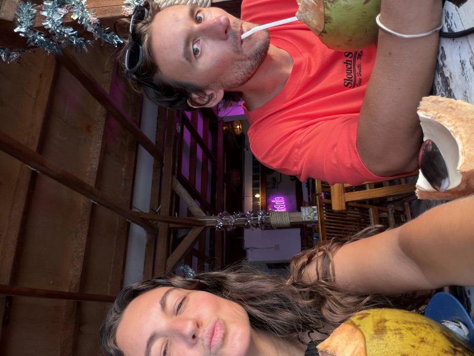 The image displays two individuals seated at a distressed white wooden table within an open-air establishment in Jimbaran, Indonesia. On the left, a female subject with long, dark, wavy hair, wearing a dark top, puckers her lips and partially closes her eyes, looking towards the camera. A whole coconut drink in a blue holder is positioned to her left on the table. On the right, a male subject with dark hair and sunglasses perched on his head, wearing a red t-shirt with visible text "Slouch S", drinks from a whole green coconut with a white straw while looking towards the camera. He wears a silver-colored bracelet on his right wrist. A split coconut shell with a spoon rests on the table in front of him. The background features a wooden beamed ceiling structure decorated with silver tinsel. Visible further back are wooden chairs and tables, a "FRIES CHILL" sign, and a pink neon sign displaying partial, inverted text, illuminated by additional yellow artificial lights.