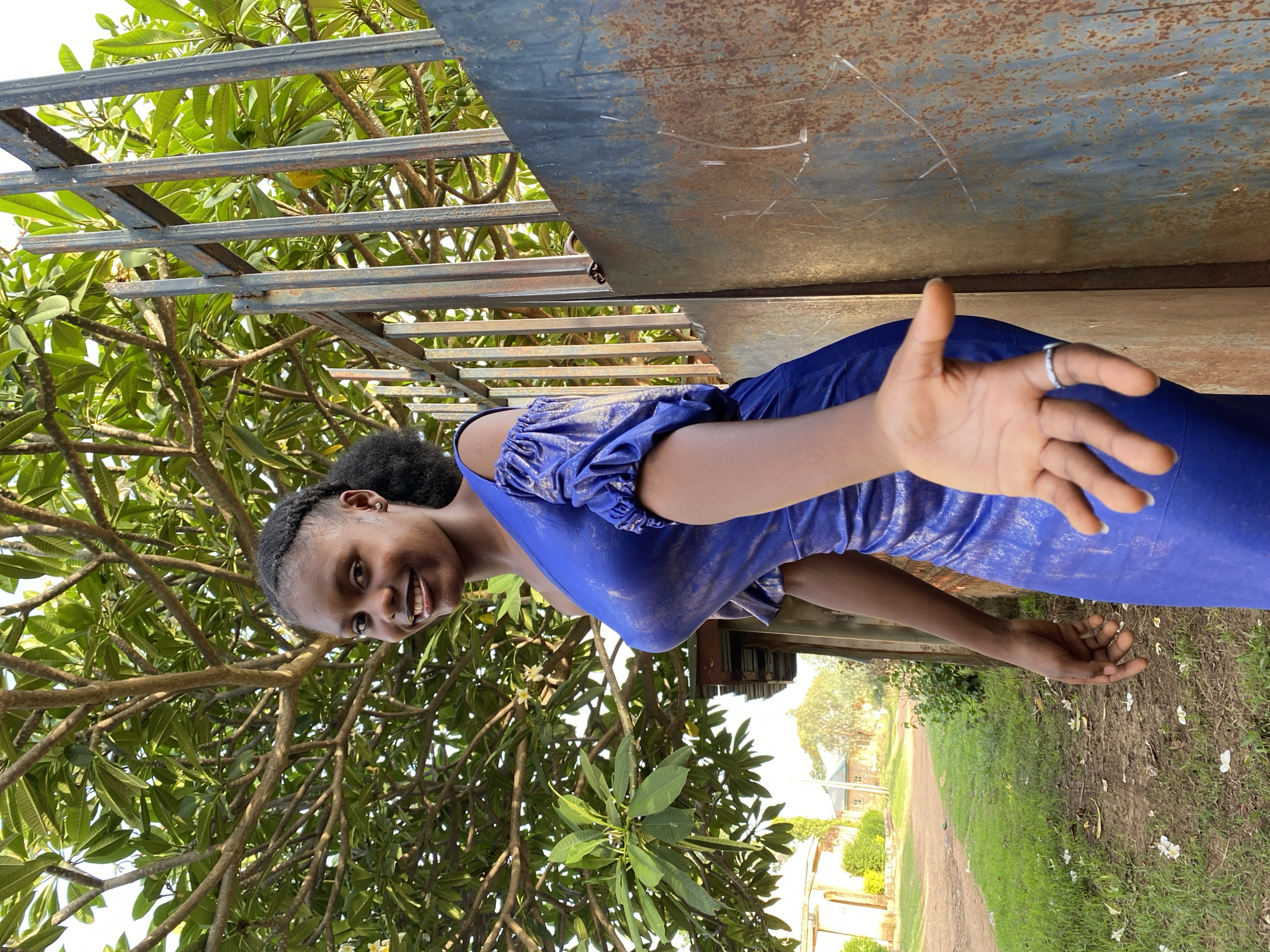 This media file features a smiling young woman outdoors in Vom, Nigeria, captured in bright daylight. She has dark skin and braided hair, and is wearing a vibrant, shiny blue dress with ruched, puffy sleeves. Her right hand is extended towards the camera, palm slightly open, revealing a silver ring on her ring finger. Her left hand is partially visible, resting against her dress.
Behind the woman, a rusty, dark grey metal structure, possibly a gate or overhead frame, spans across the upper portion of the background. Lush green foliage from a large tree with prominent branches frames the left side and top of the image. The right side of the background shows a dirt path or road curving into the distance, flanked by green grass and white flower petals scattered on the ground. Several distant buildings are visible beyond the path. The overall mood is cheerful, conveyed by the woman's broad smile and inviting gesture. There is no visible text.