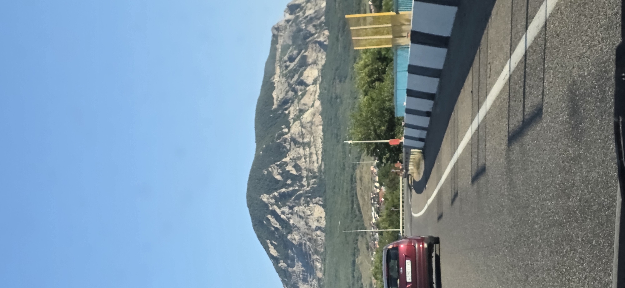The image depicts a roadside view with a mountain in the background. In the foreground, a red car is driving on an asphalt road. To the right of the road, there is a barrier made of alternating black and white sections, and beyond that, a blue structure and a yellow object are visible. A few trees are scattered throughout the landscape, and in the middle ground, a village with houses can be seen nestled at the foot of the mountain. The mountain itself is large, with rocky outcrops and green vegetation covering its slopes. The sky is clear and blue. The location is identified as Zmeyka, Russia, suggesting this is a scene from a highway leading towards or away from the mountain.