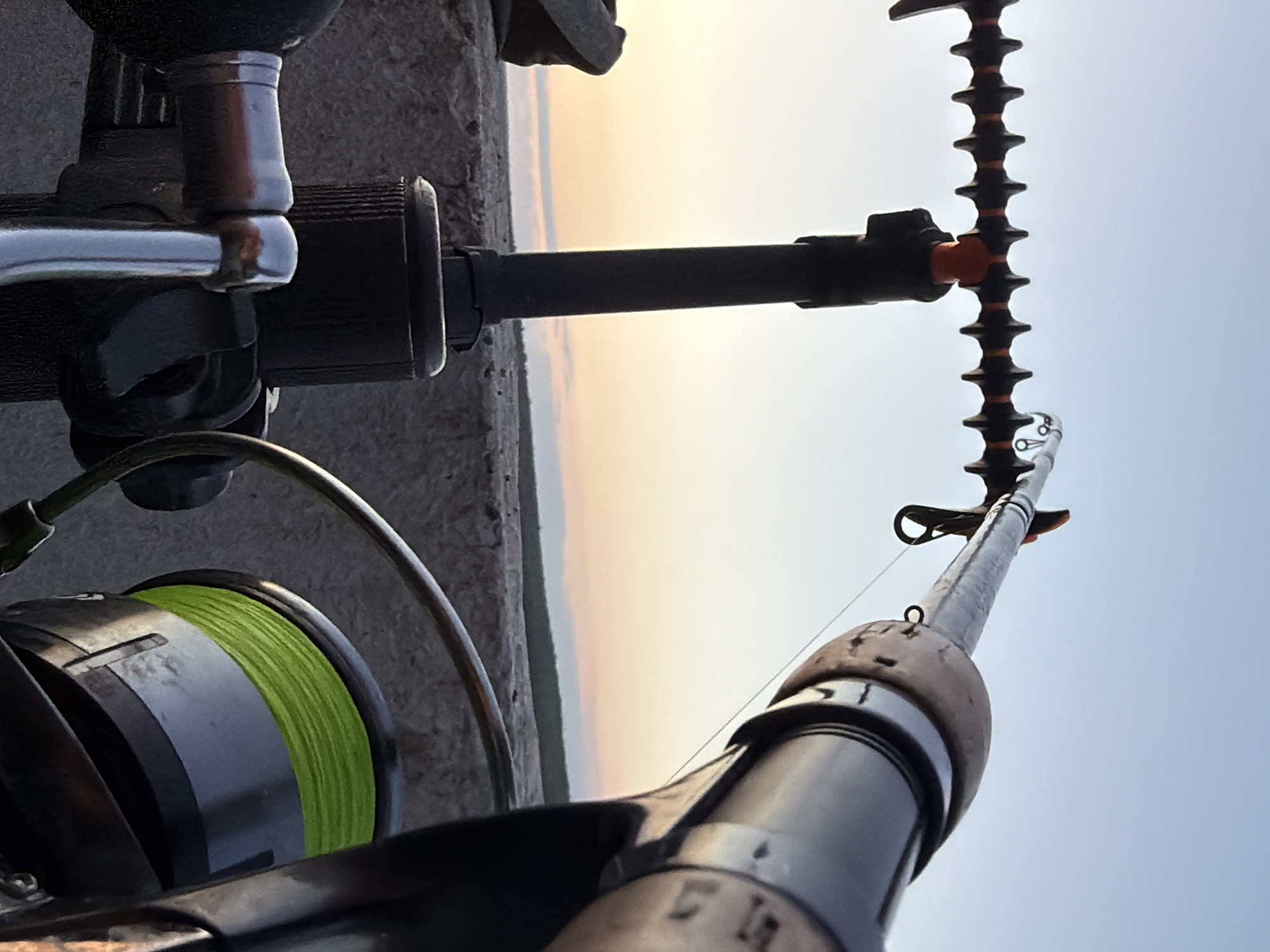 The image shows a close-up, low-angle view of fishing equipment set up on what appears to be a concrete embankment. The foreground is dominated by two fishing rods mounted on black rod pods. One rod, on the right, is in focus, revealing a thick, dark handle with a wooden grip, a reel with bright green fishing line, and a fishing line extending upwards. The other rod, on the left, is less in focus but shows part of its reel handle and a dark rod.
In the background, the sky transitions from a pale blue at the top to a soft gradient of peach and pink near the horizon, indicating either sunrise or sunset. A thin, dark line of vegetation or trees is visible on the horizon, separating the embankment from the sky. The embankment itself is rough and textured, appearing to be made of concrete or stone. No people are visible in the frame. The scene suggests a quiet fishing spot, possibly overlooking a body of water, though the water itself is not depicted. The specific location in Chaikovsky, Russia, is not visually identifiable from the provided image.