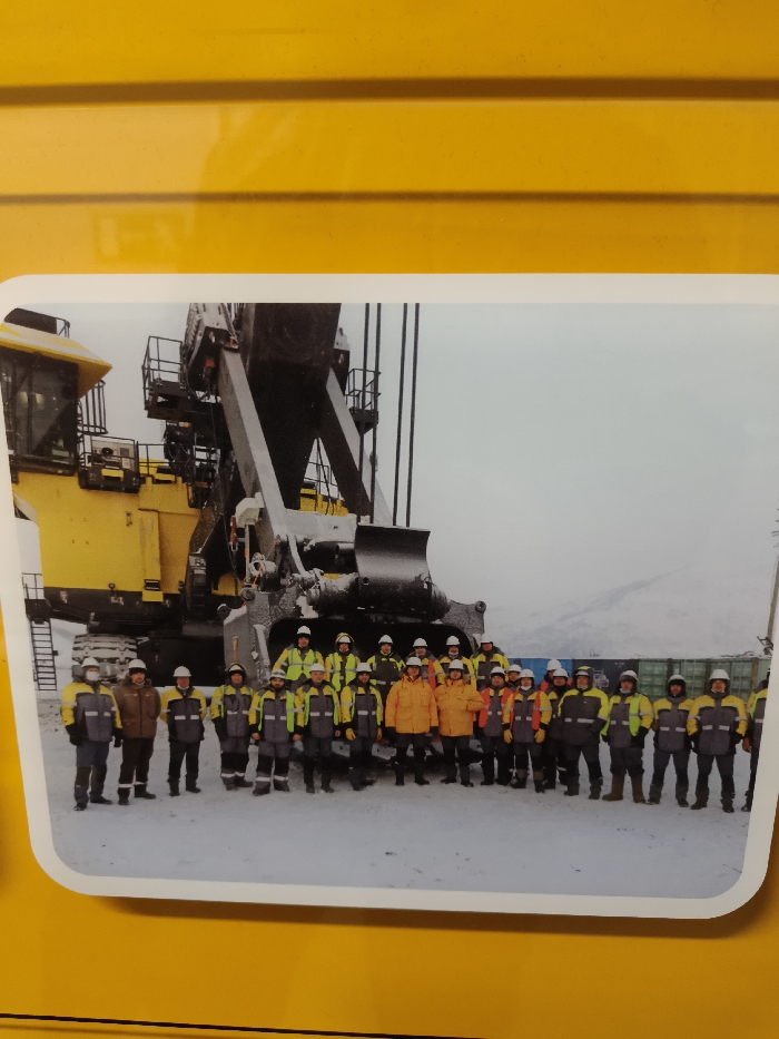 A photograph captures approximately 22 individuals standing in a snowy outdoor environment, posing in two rows for a group portrait. The scene is set in the city of Im. Gastello, Russia. The group is positioned in front of a large yellow and grey tracked mining excavator, which dominates the left and central background. The excavator's substantial bucket is visible, showing snow accumulation. All individuals are uniformly dressed in cold-weather work attire, consisting of high-visibility yellow or orange jackets, grey trousers, and most wear white hard hats. Some individuals are also wearing face coverings. The ground is covered with snow, and in the distance, a range of snow-covered mountains is visible under an overcast, light grey sky. To the far right of the frame, several green cargo containers or rail cars are partially visible.