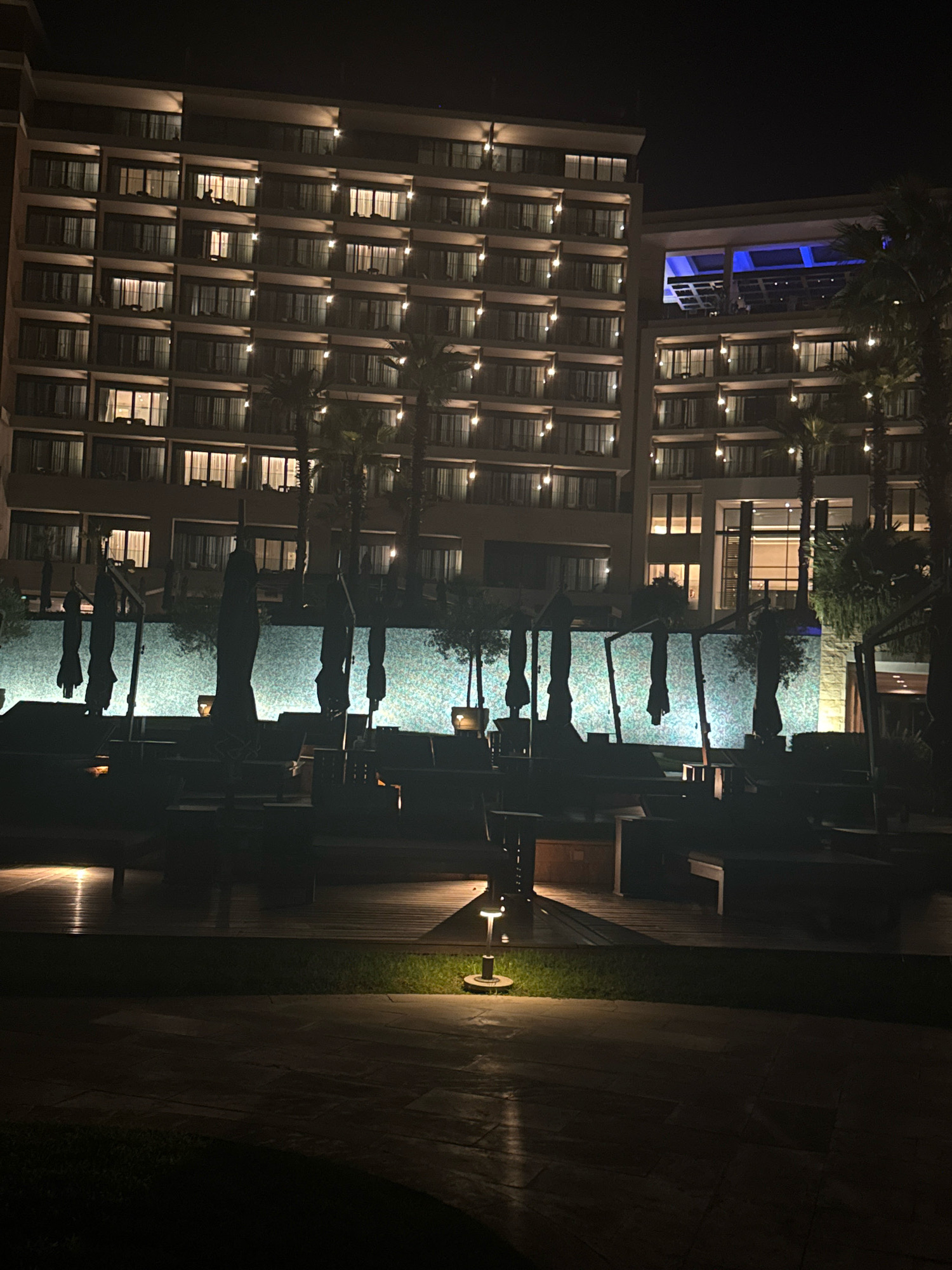 At night, the exterior of a large hotel building is illuminated. The hotel has multiple floors with many windows, some of which are lit from within. In front of the hotel, there is a swimming pool area with lounge chairs and umbrellas. The pool is lit with a blueish light, and the surrounding area is paved with wood and stone. There are palm trees visible around the pool area. The scene appears to be part of a resort or hotel in Agios Tychona Community, Cyprus.