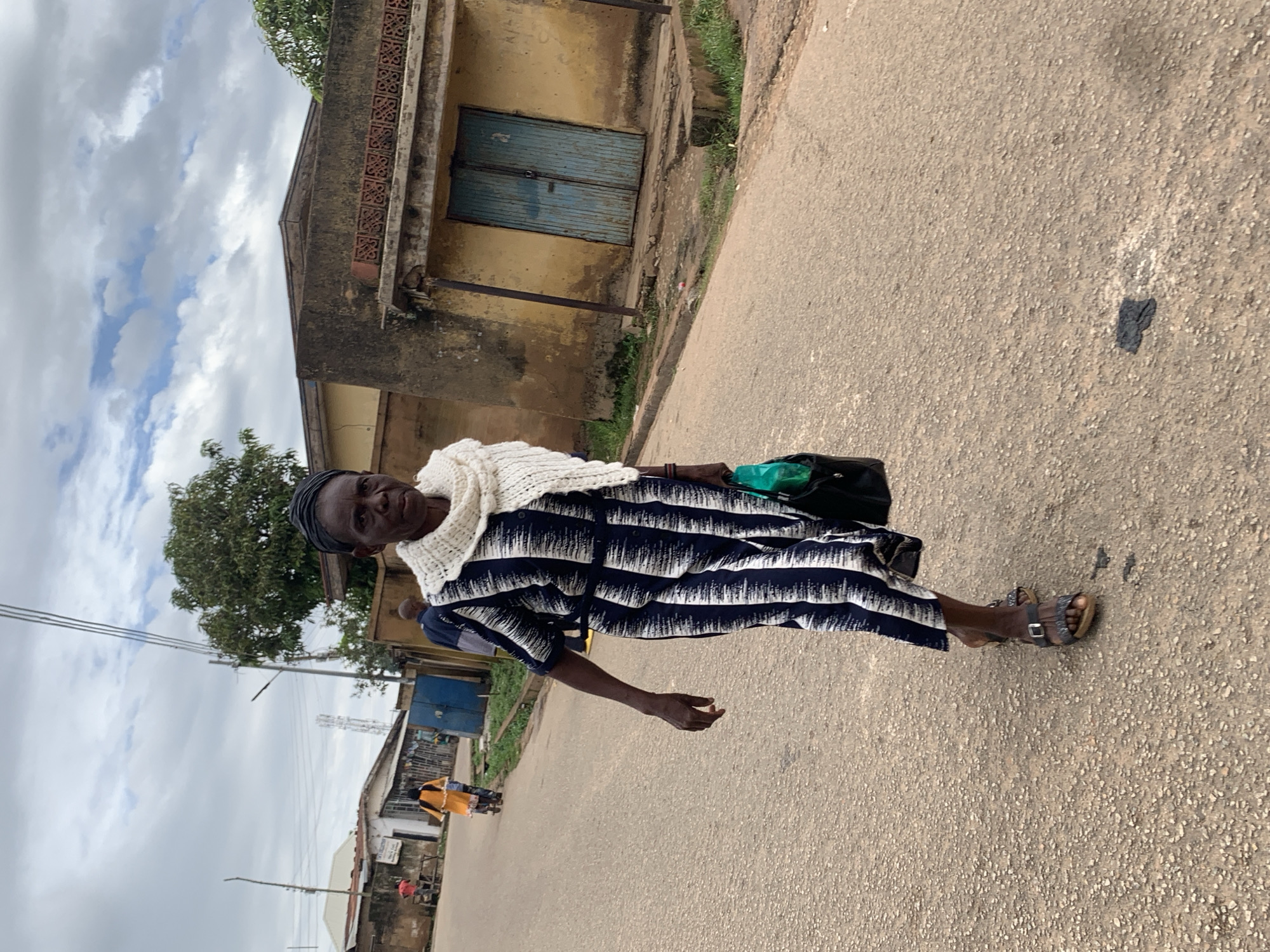 A Black woman, appearing to be in her late 50s or 60s, walks towards the camera on a paved road in Jos, Nigeria. She is wearing a dark blue and white patterned dress, a thick white knitted scarf around her neck, and dark sandals. She carries a black handbag with a green item visible inside, and her left hand is resting on the bag. Her hair is covered by a dark headscarf.
In the background, a yellow building with a brown trim and blue double doors is visible on the right. A tree with green foliage stands behind the building. Further in the distance, to the left of the frame, several other buildings and structures are present, including what appears to be a red-roofed building and some antennas. Other individuals and a person on a motorbike are faintly visible further down the road, moving away from the viewer. The sky is overcast with grey and white clouds. The road surface is a rough asphalt with small pebbles.