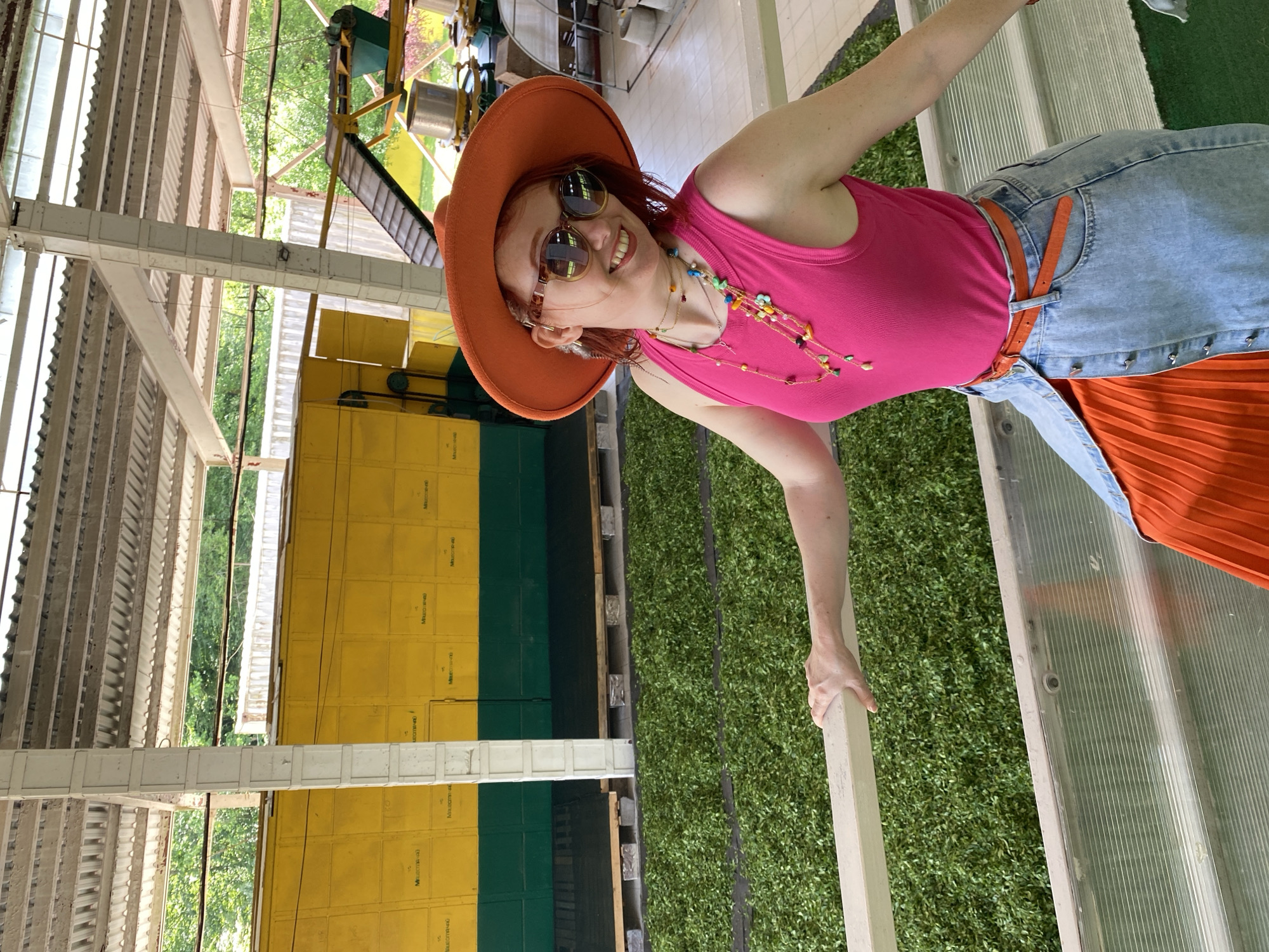 A woman in a pink tank top, blue skirt, and orange hat is posing indoors. She is wearing sunglasses and a multi-colored necklace. The indoor space features a green floor, a yellow and green wall, and a white metal structure. In the background, machinery is visible, along with green foliage. The setting appears to be in Izmaylovka, Russia.
