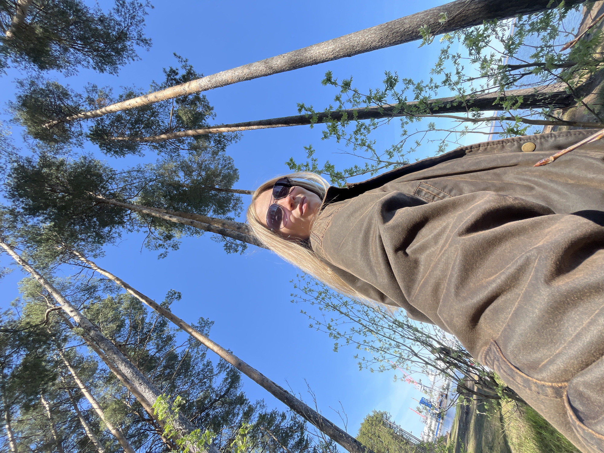 The photograph, taken from a low-angle perspective, shows a blonde woman wearing sunglasses and a brown jacket in an outdoor setting dominated by tall trees against a bright blue sky. The location is suggested to be Chaikovsky, Russia, based on distant structures that appear to be a shipyard or industrial complex, visible through foliage in the background. The woman is positioned in the foreground, with the trees towering above and around her. The trees are interspersed with younger trees and some budding foliage, suggesting spring. The lighting is bright, creating strong shadows, and the overall environment suggests a natural area with some human presence nearby.