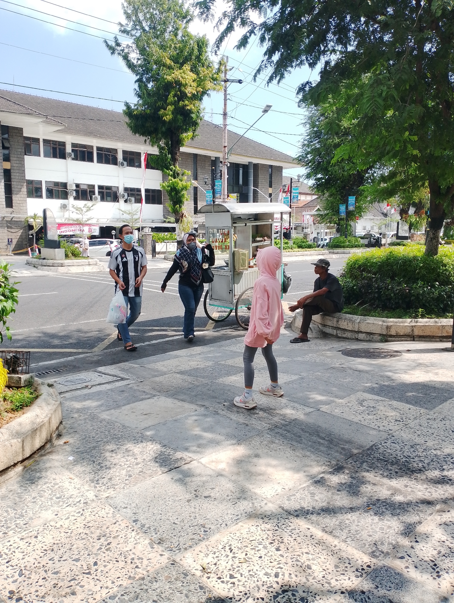The image captures a street scene in Yogyakarta, Indonesia, with a focus on the pedestrian area and a road. In the foreground, a young person wearing a pink hooded sweatshirt and grey leggings stands facing away from the camera, with their shadow cast on the patterned pavement. To the left, two adults are walking across the road. The man, wearing a black and white striped jersey and blue jeans, carries a white plastic bag. He is wearing a light blue face mask. Beside him, a woman in a blue patterned hijab and blue jeans also wears a face mask. Behind them, a food cart with a white canopy is parked near the sidewalk. Further back, a man in a dark t-shirt and a cap is seated on a low stone wall, partially shaded by a large tree. The background shows buildings with gray stone facades and white upper stories, typical of institutional architecture in the city, with trees and some street furniture visible. Electrical wires and streetlights are also present, indicating an urban setting. The lighting suggests it is daytime, likely mid-morning or afternoon, with clear skies.
