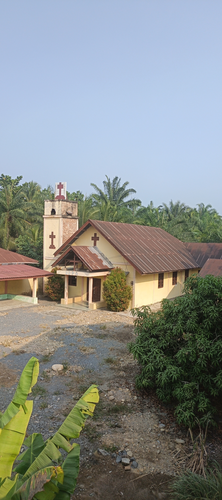 The image depicts a view of a small, modest church building situated in a rural or semi-rural area. The church is painted yellow with a rusty, corrugated metal roof. It has a prominent bell tower with crosses on its upper section and a smaller cross on the side of the main building. The entrance features a covered porch with two wooden doors. Several green bushes are planted around the entrance.
The church grounds are covered with gravel and some scattered stones. To the left of the main building, there is an adjacent structure with a similar metal roof. The background is dominated by lush green palm trees, indicating a tropical or subtropical environment. In the foreground, large green banana leaves are visible, suggesting a garden or natural vegetation. The sky is clear and pale blue, indicating daytime.
The sign above the entrance reads "GBPA." The overall scene suggests a quiet, possibly remote location, consistent with a village setting in Indonesia. The image is taken from a slightly elevated vantage point, looking down towards the church.